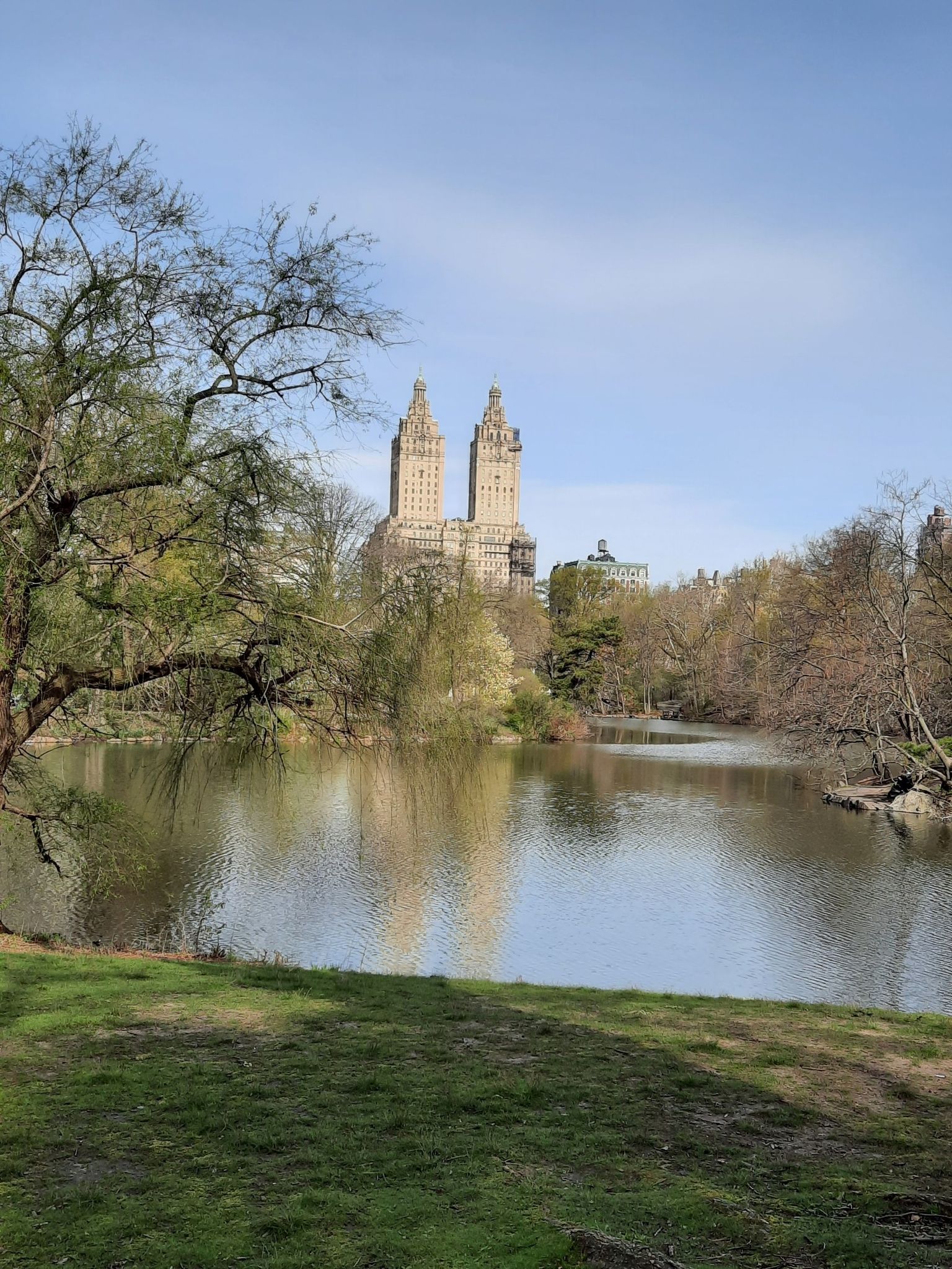 Lake reflecting twin buildings, bare and green trees, bright sky. - View of the San Remo Towers – Bethesda Fountain and Terrace loop from 59th Street