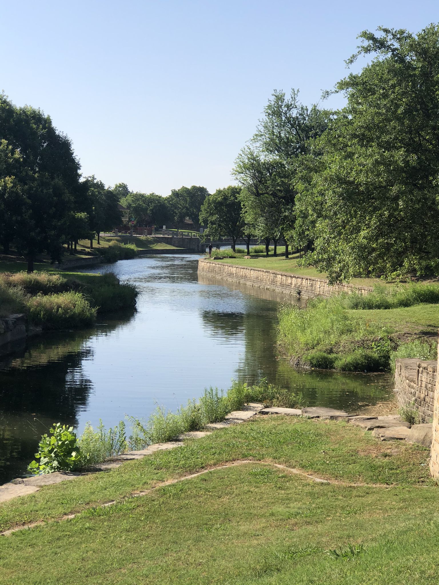 A calm river flows between grassy banks and lush green trees on a sunny day. - Chisholm Trail