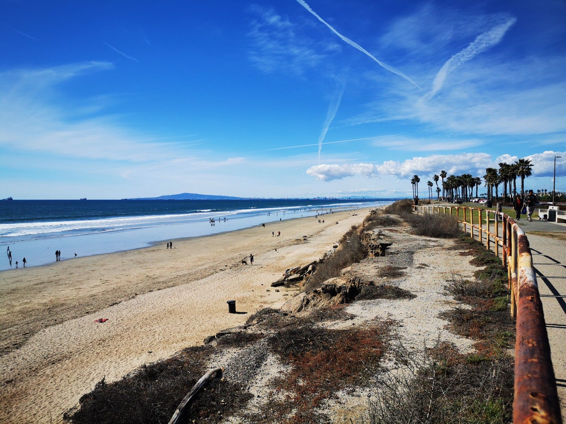 Expansive sandy beach with ocean waves, distant people, and a coastal path. Bright day. - Huntington Beach Bike Trail