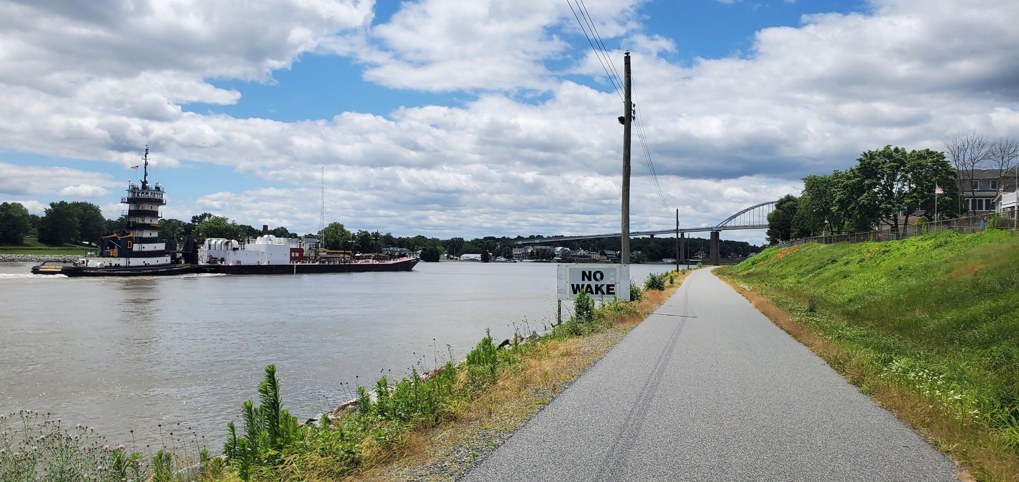 A tugboat on a river next to a paved path and a "NO WAKE" sign. - C&D Canal Trail