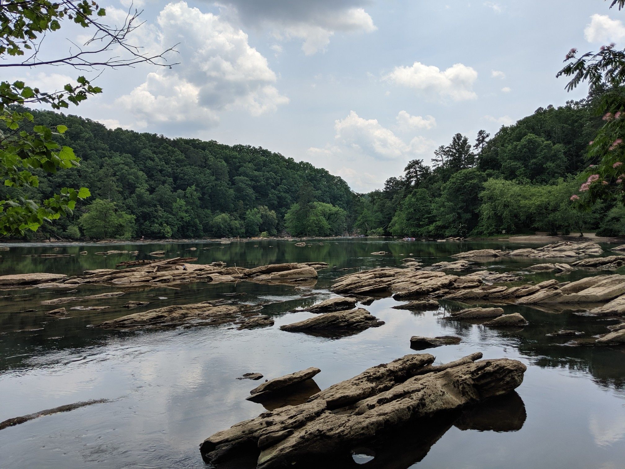 River with exposed rocks, dense green forest, and cloudy sky. - East Palisades Trail