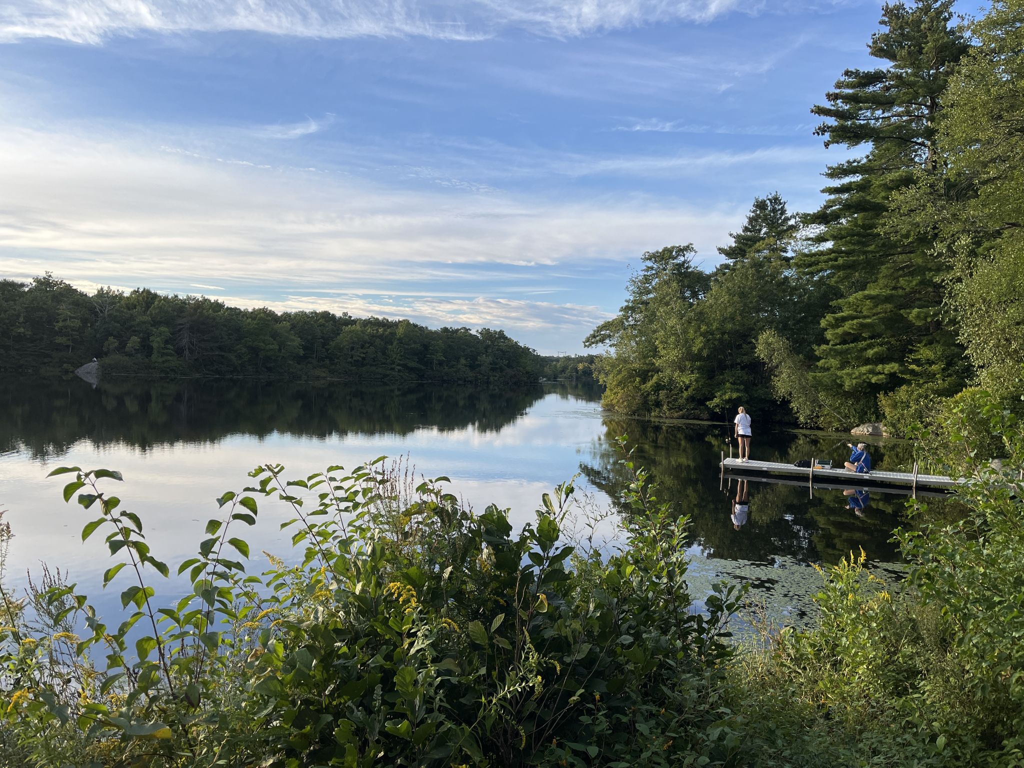 Two people on a dock by a calm lake surrounded by green trees. - Ames Nowell Loop