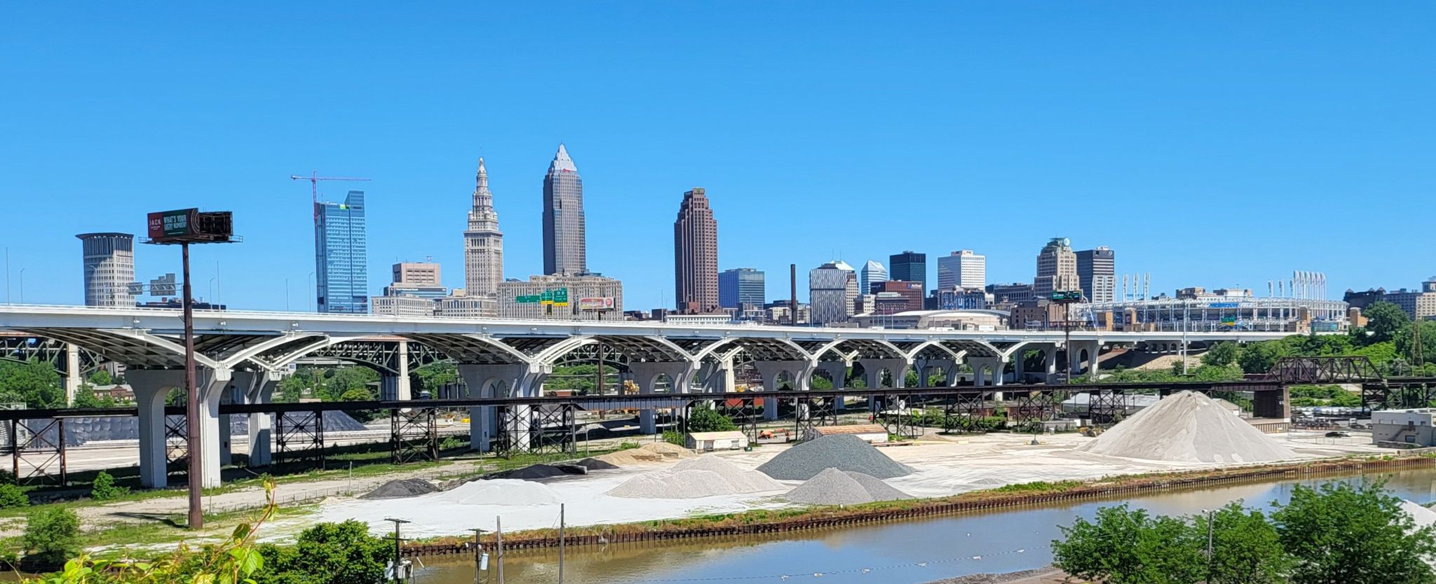 City skyline behind a large arched bridge, river, and material piles. - Ohio & Erie Canal Reservation – West Side Market loop from West 65th–Lorain (Eco-Village)