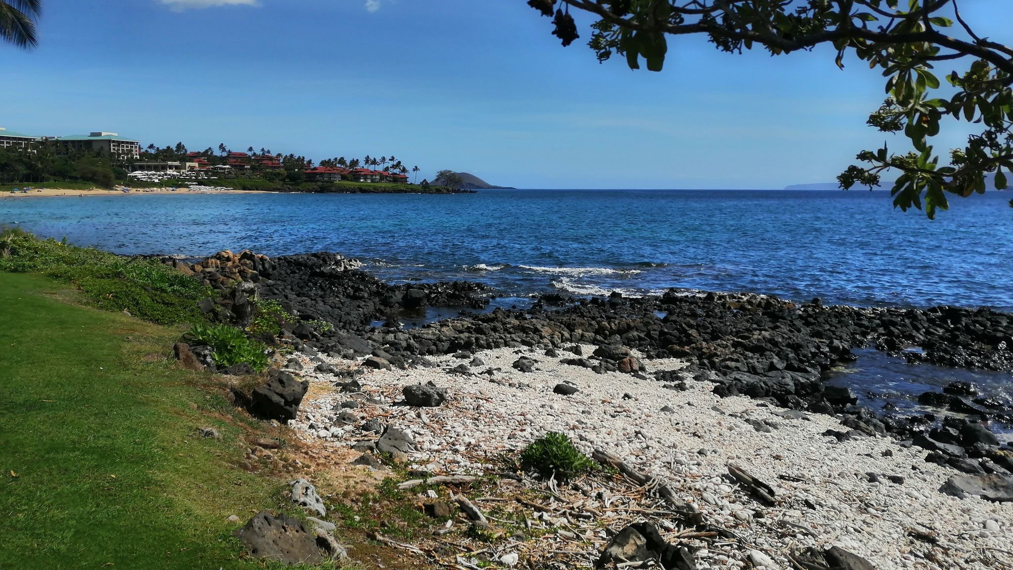 Rocky and sandy beach with green grass, blue ocean, and distant land. - Keawakapu Beach – Keawakapu Beach loop from Kamaole Shopping Center (northbound)