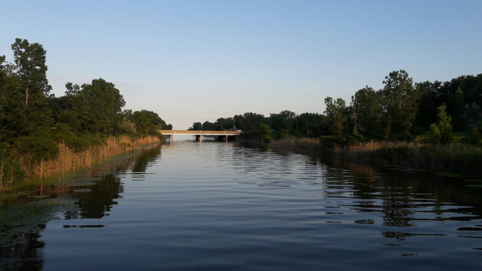 Calm river flowing under a distant bridge, lined by trees at sunset. - Lake St. Clair Metropark Beach loop from 15 Mile / Maplegrove (EB)