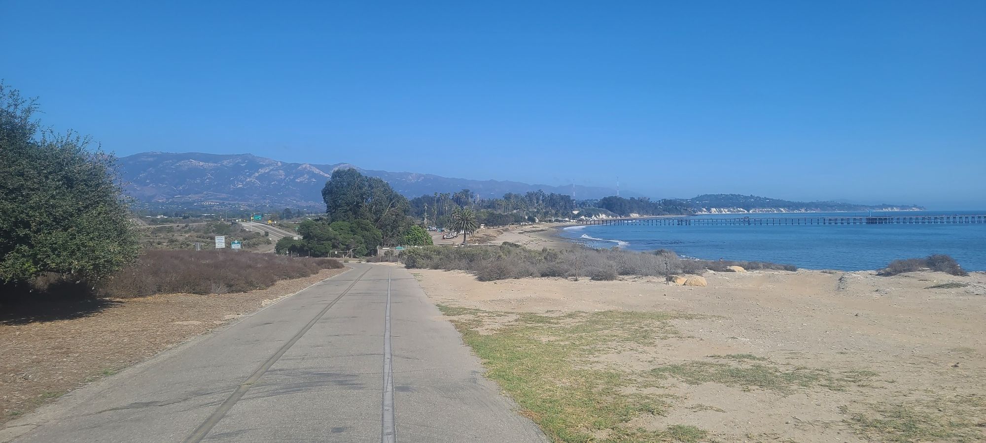 Paved road by a sandy beach and ocean with distant mountains under clear sky. - University of California to Goleta Beach Park run
