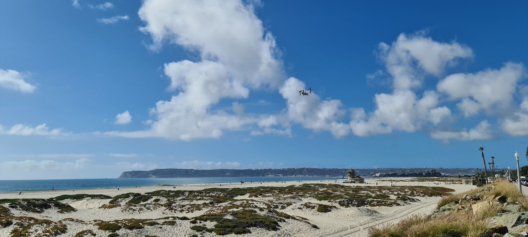 Wide sandy beach, ocean, distant land, and a helicopter under a bright, cloudy sky. - Coronado Beach