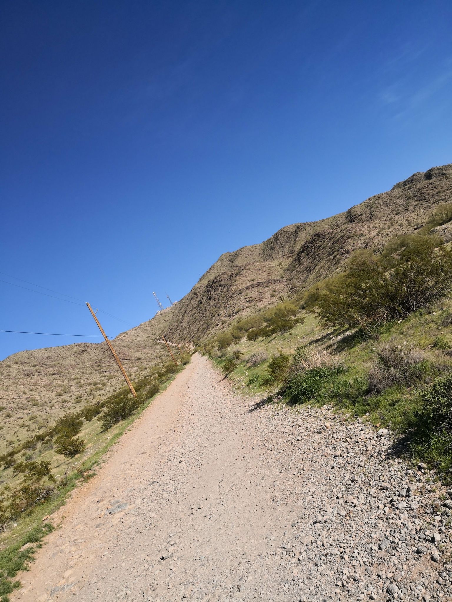 A dirt path ascends a steep, rocky, sunlit desert hillside under blue sky. - Hiking loop from Casa Grande 2 Electronic Site