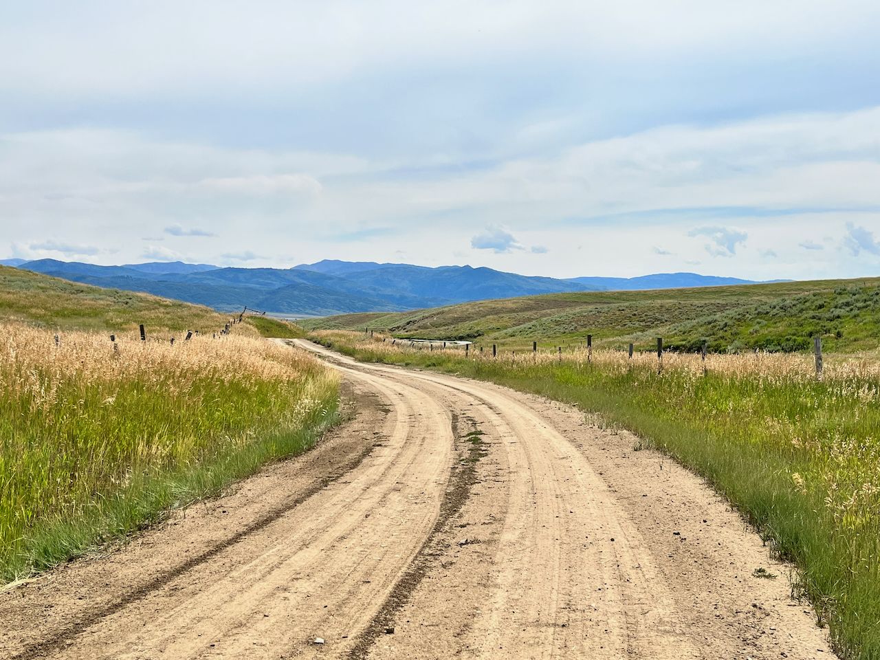 Winding dirt road through golden grassy hills with distant mountains under cloudy sky. - Elk River – Yampa riverside park loop from Steamboat 2