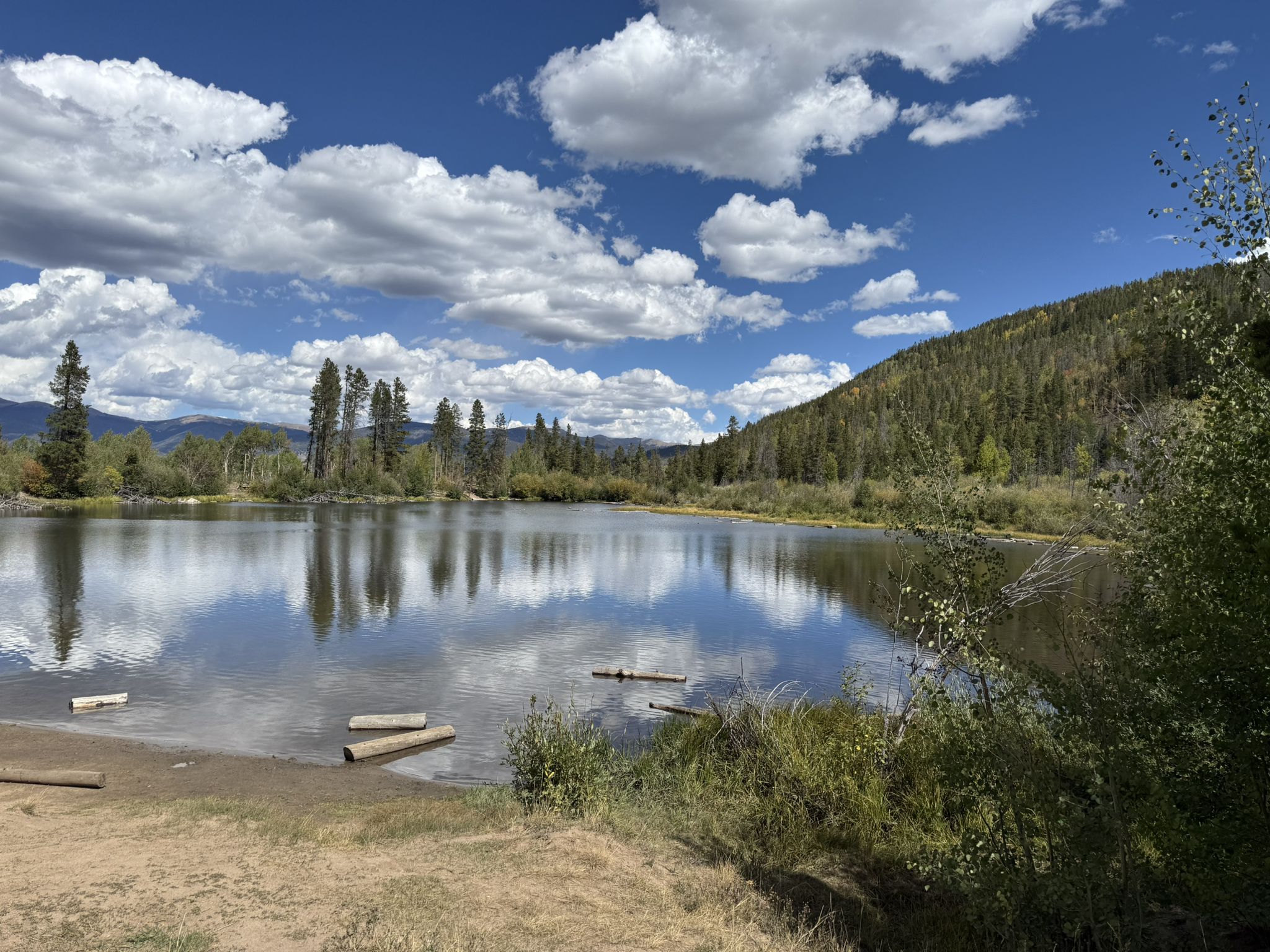 Serene lake reflecting blue sky and clouds, bordered by lush green forest. - Peaks Trail