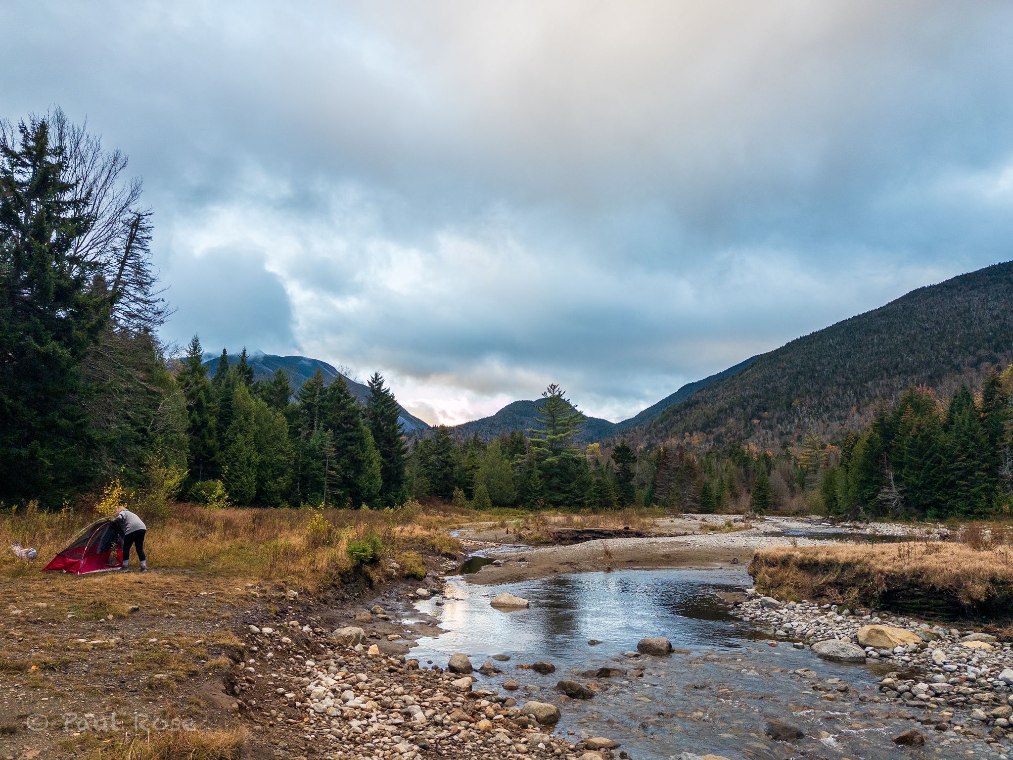 A person setting up a red tent by a river with forested mountains under cloudy skies. - Mount Marcy – Indian Falls Lookout loop from South Meadows Parking Area