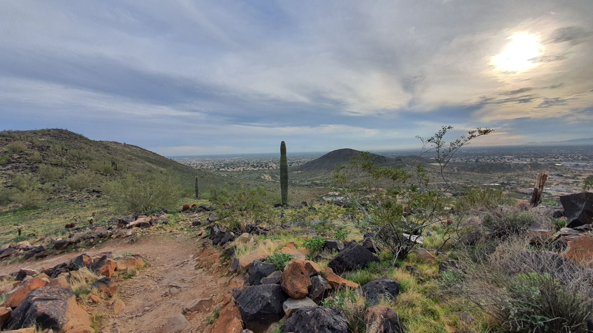 Rocky desert path leading to distant city under cloudy sky with sun. - Thunderbird Park Three Peaks Loop Trail