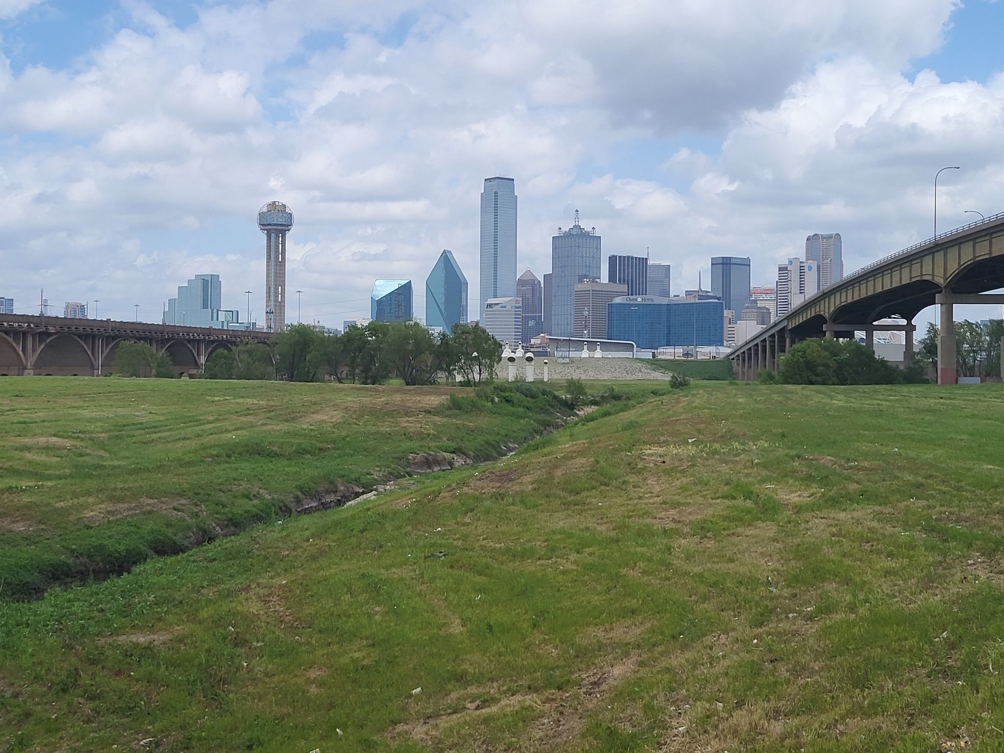 Grassy field with stream, bridges, and a city skyline under cloudy sky. - Dallas Morning News loop from Kay Bailey Hutchinson Convention Center