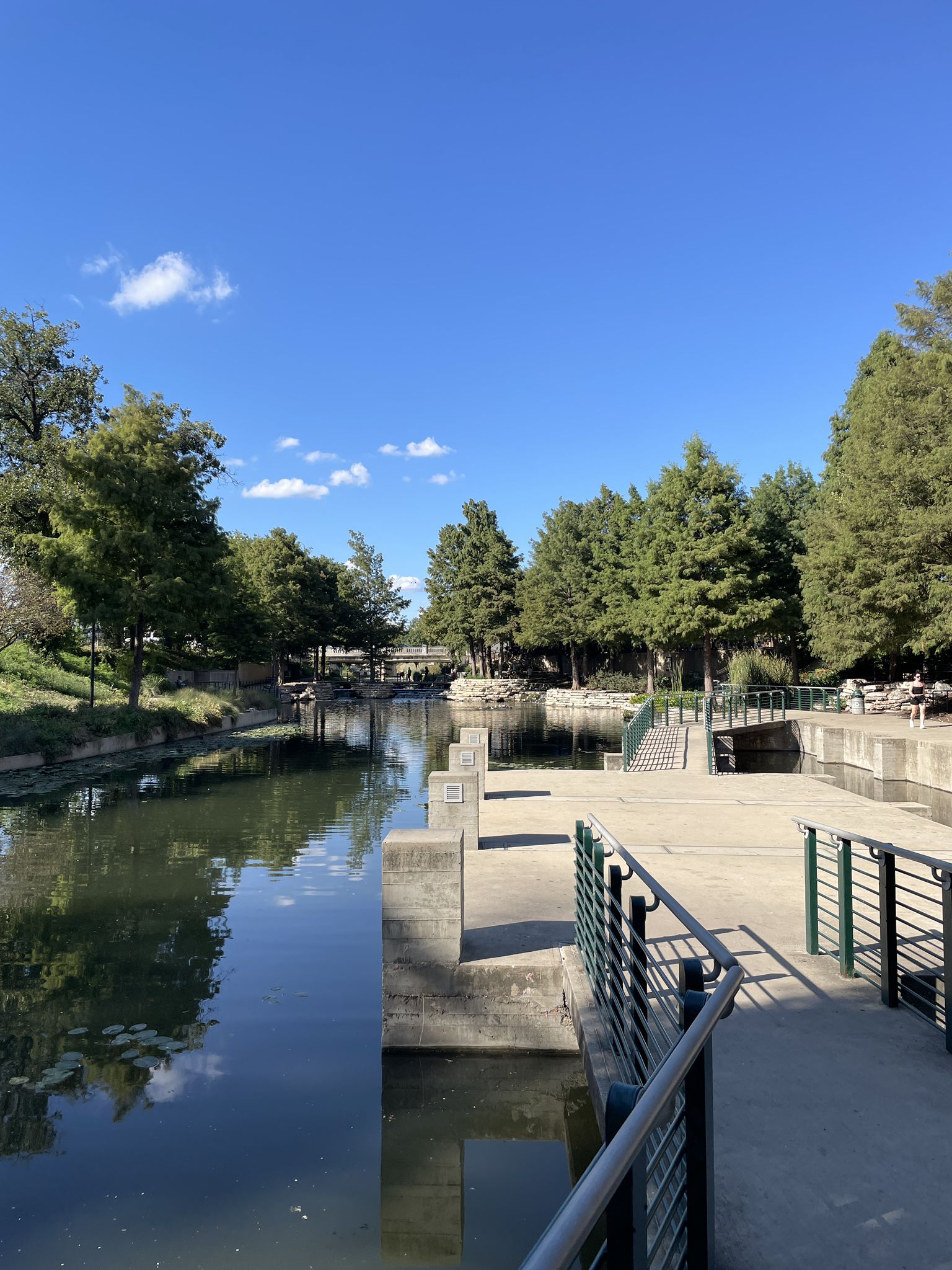 River with concrete platforms, railings, and evergreen trees under blue sky. - Alamo Mills Dam loop from Trinity University