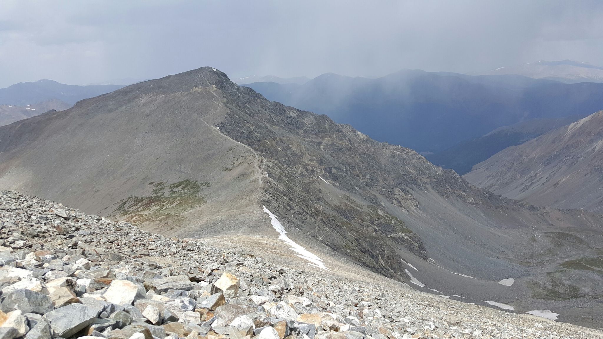 A steep, rocky mountain ridge with snow patches under a cloudy sky. - Grays Peak Trail