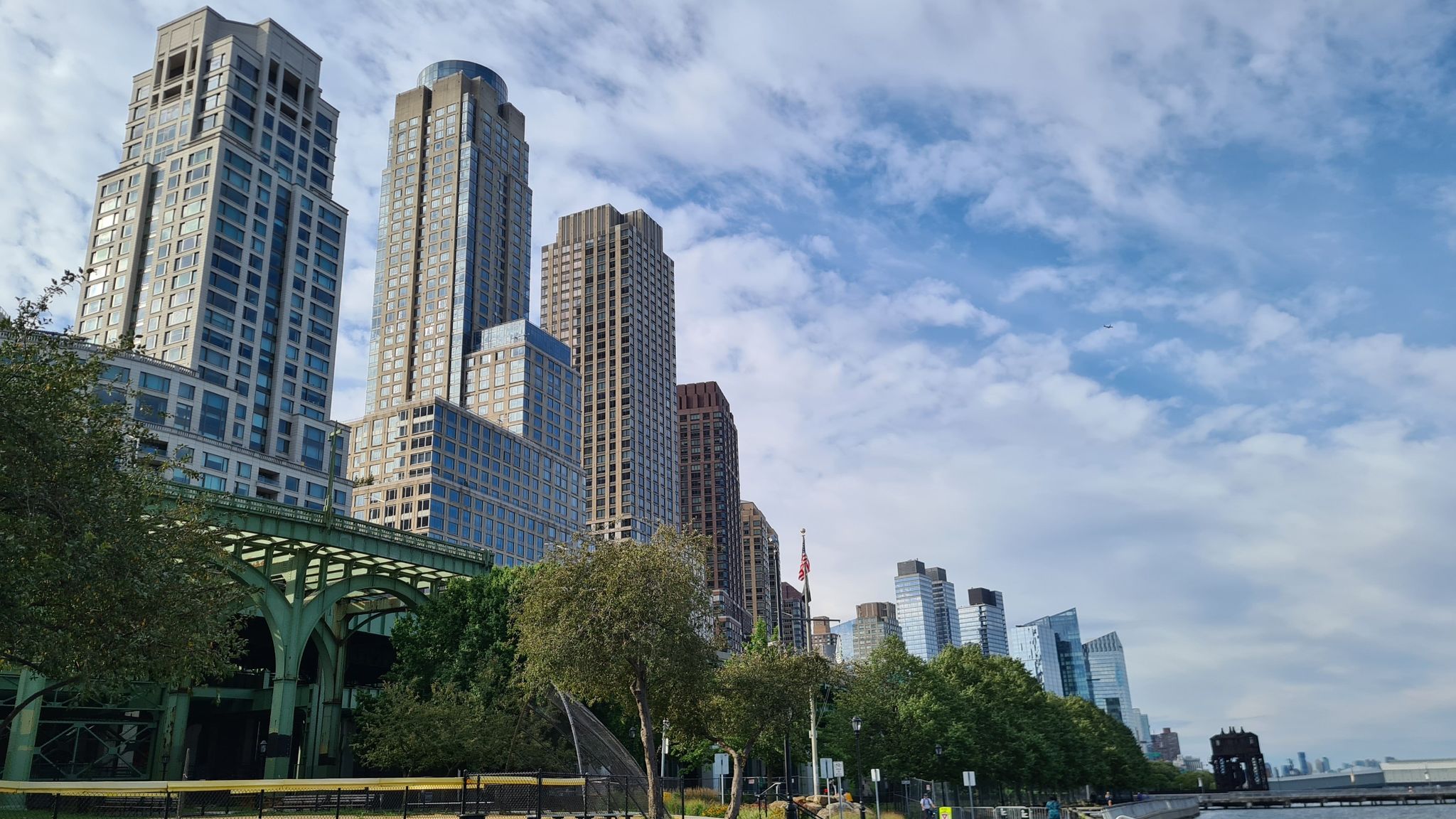 Tall city buildings, green trees, and a bridge under a cloudy blue sky. - Manhattan School of Music loop from 125th Street