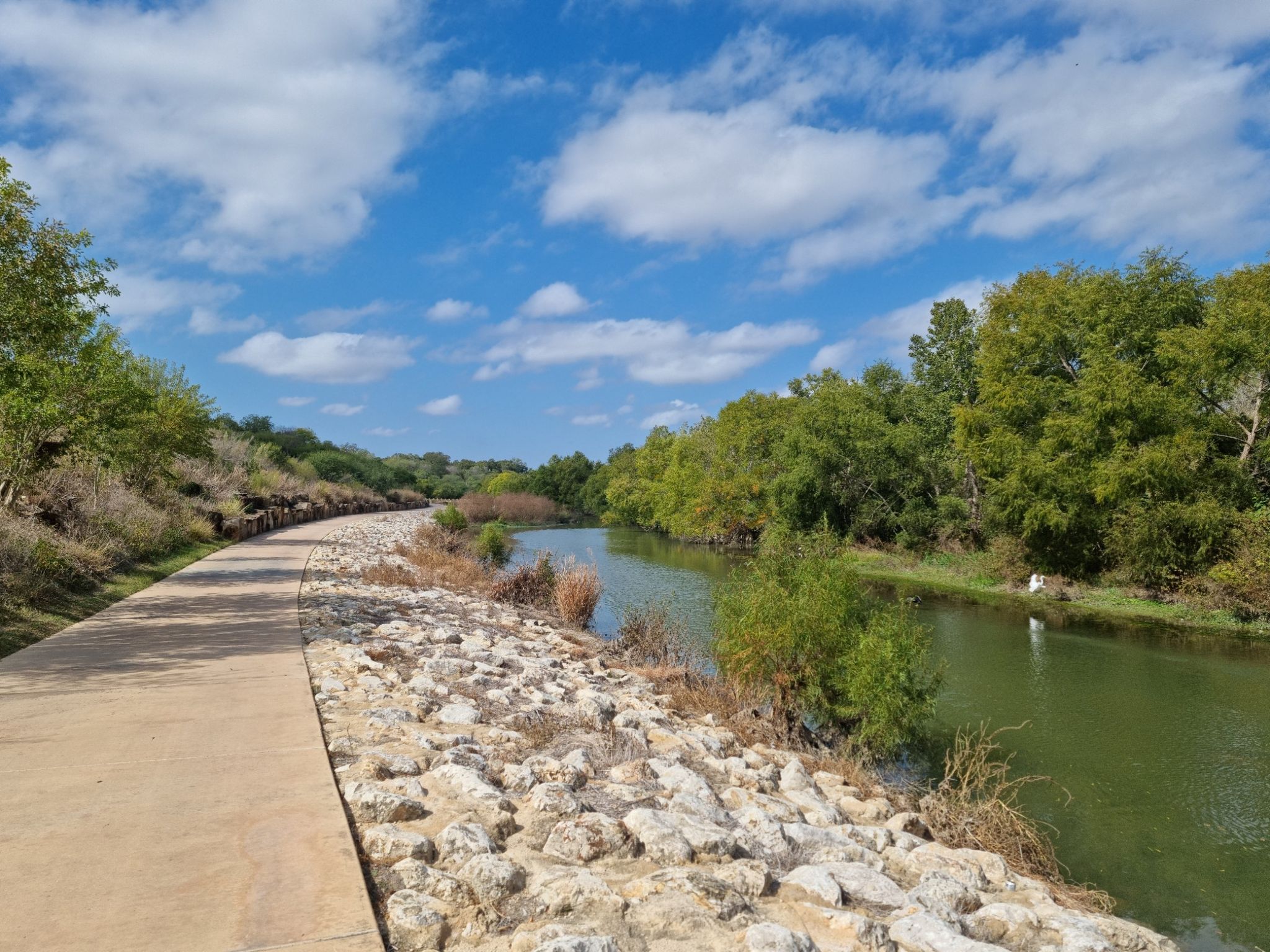Paved path alongside a river with lush green trees under a blue sky. - River Walk Mission Reach