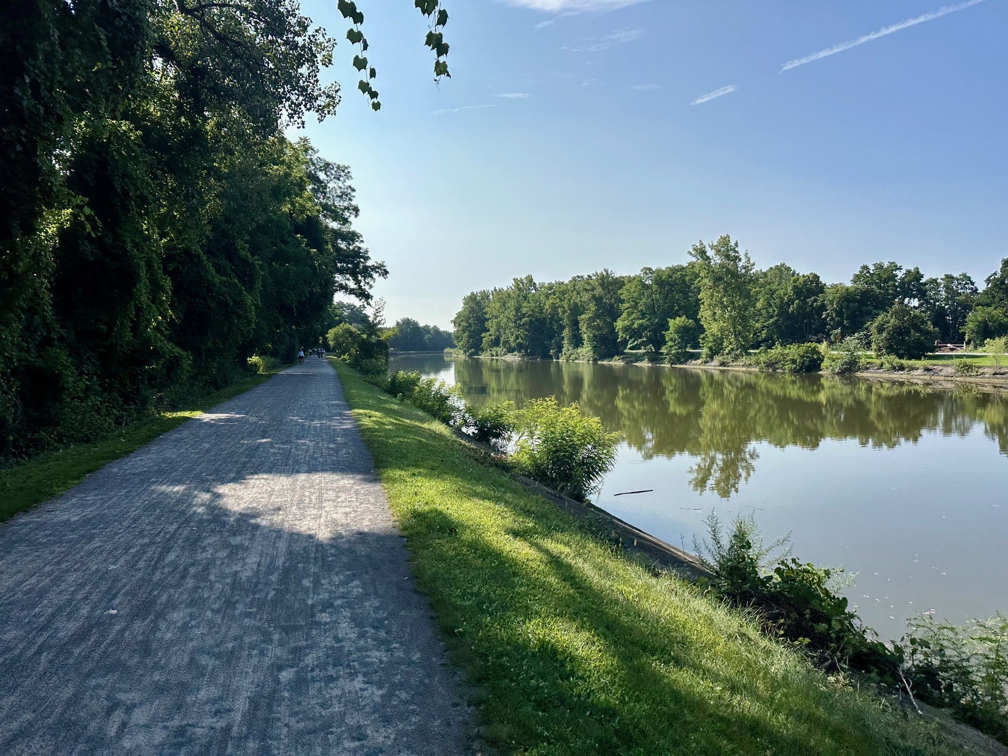 A paved path next to a calm river reflecting trees on a sunny day. - Erie Canalway Trail – Pittsford loop from Village of Fairport