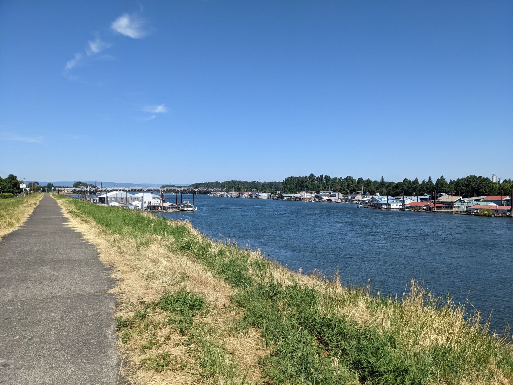Paved path alongside a river with houseboats and trees under a clear sky. - Columbia River Loop