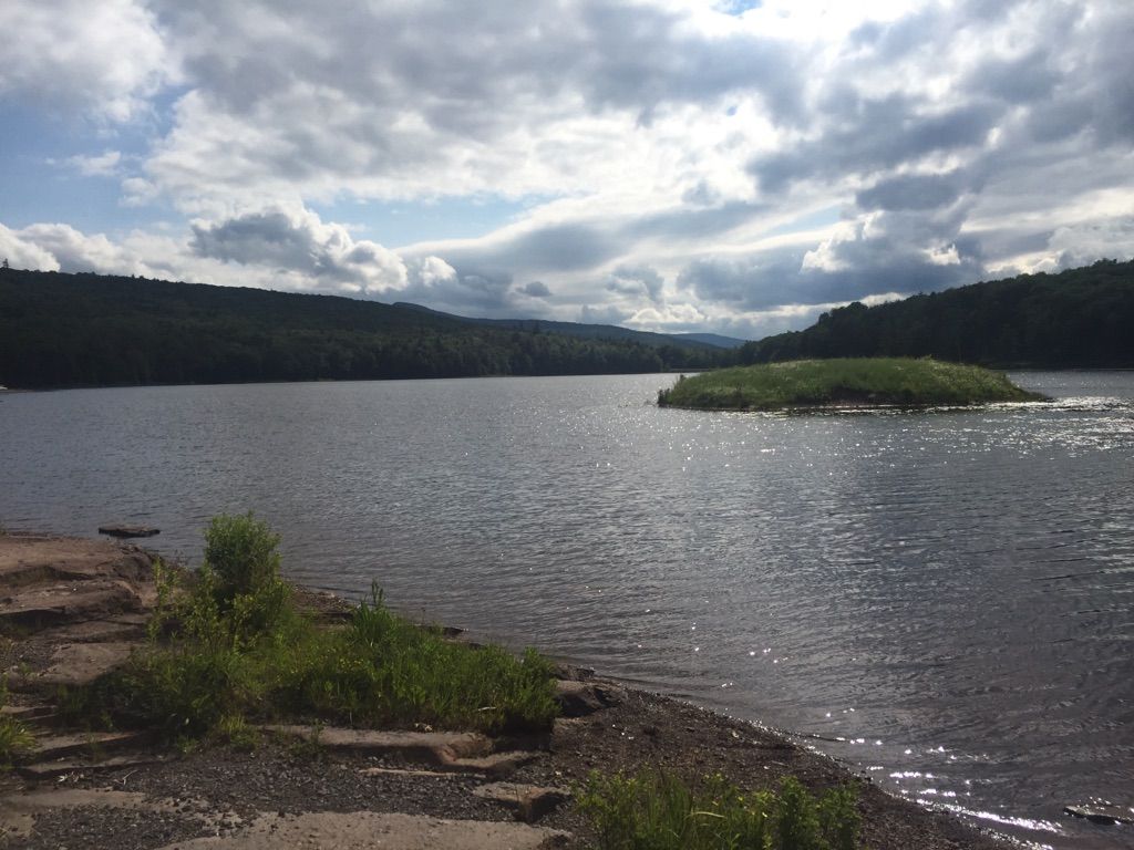 Lake with small island, forested hills, and cloudy sky over rocky shore. - North Lake South Lake Trail