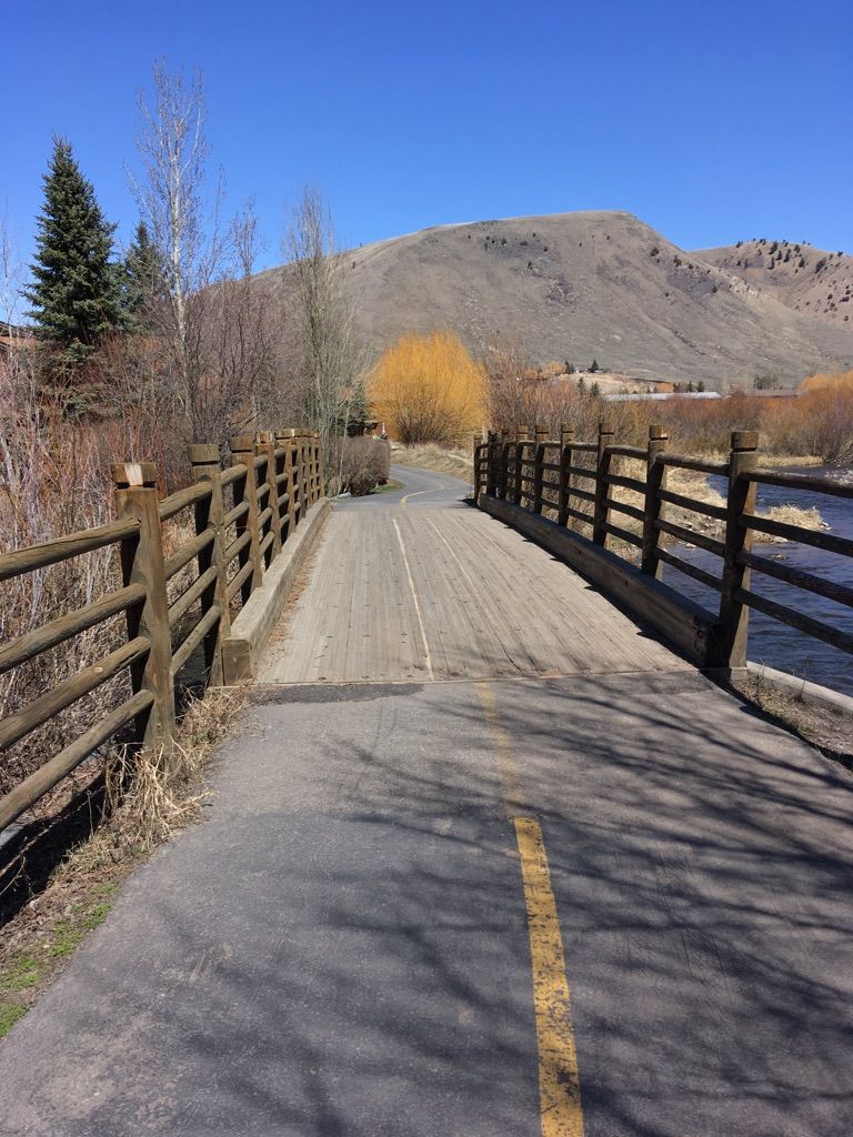 Paved path leading onto a wooden bridge over water, mountains beyond. - Snow King Mountain – Sink or Swim Trail loop from Maverick, Broadway and Huff