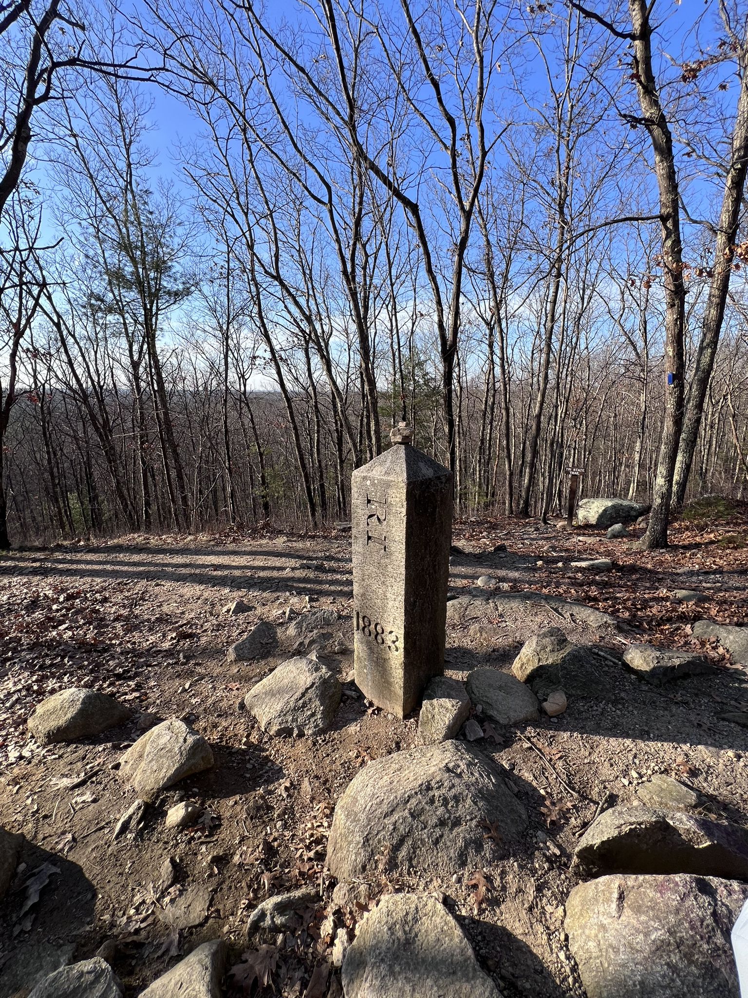 A stone marker stands on a rocky forest floor under a clear sky. - Douglas State Forest – Tri-State-Marker Loop