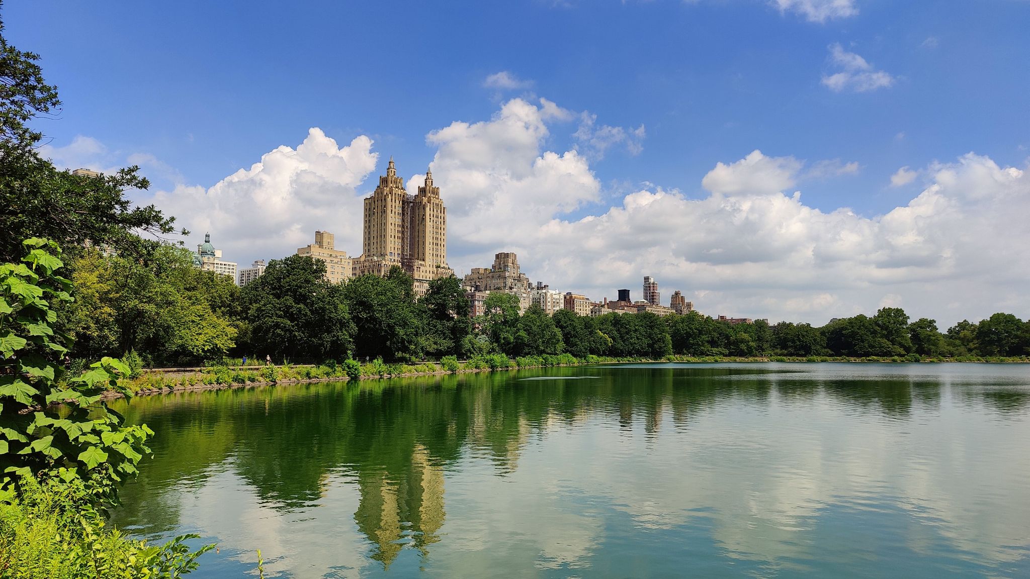 Lake reflecting buildings and trees under a bright, cloudy sky. - Central Park South – Frisbee Hill loop from 57th Street