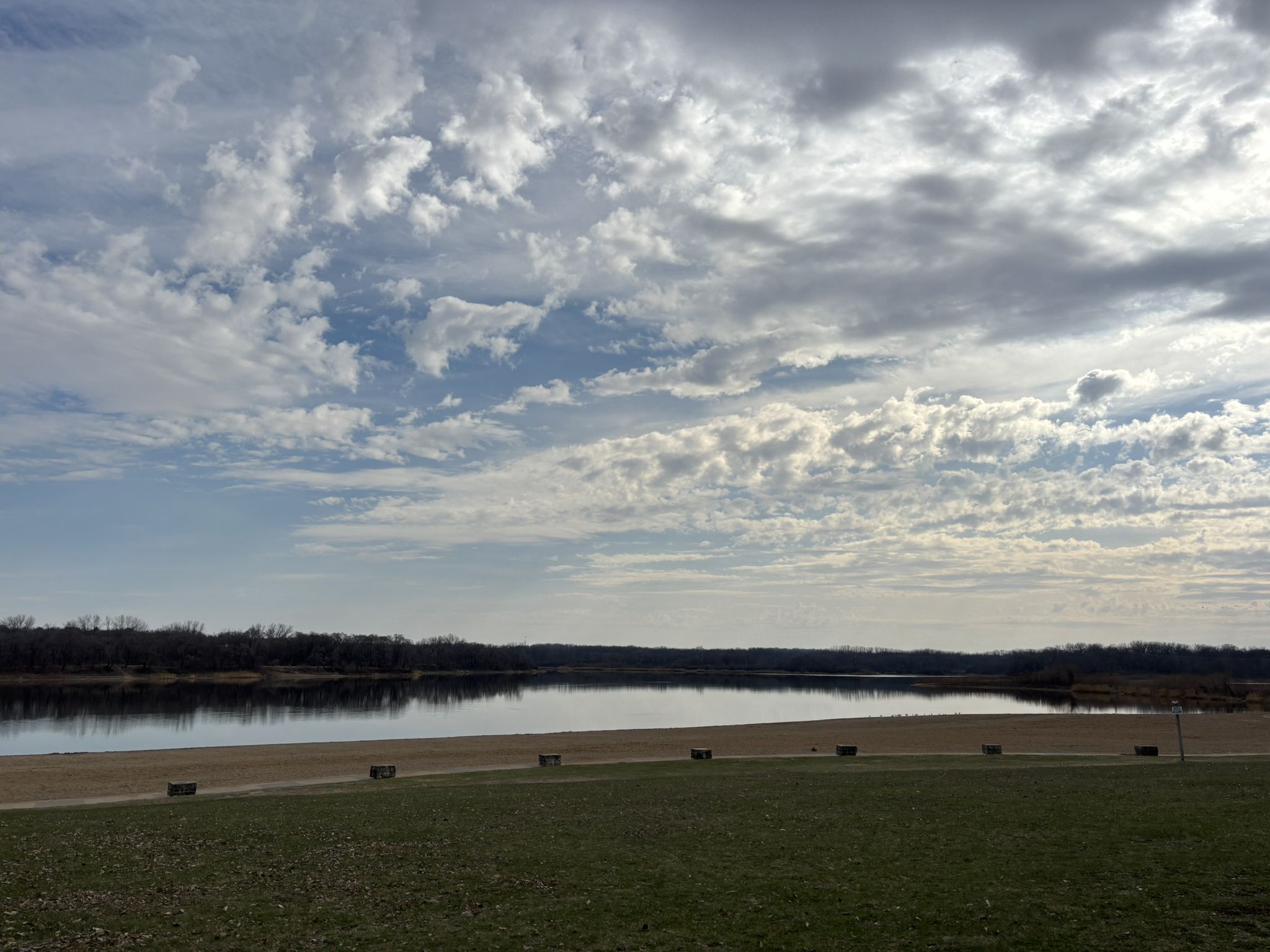 Calm lake with sandy shore under a cloudy sky, bordered by distant trees. - Raccoon River Park Trail