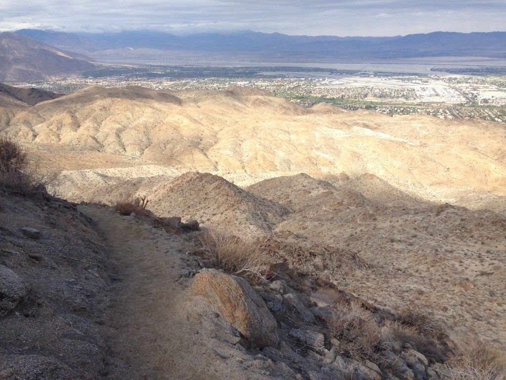 A desert trail winds through arid mountains overlooking a distant city. - Goat Trails to Murray Hill