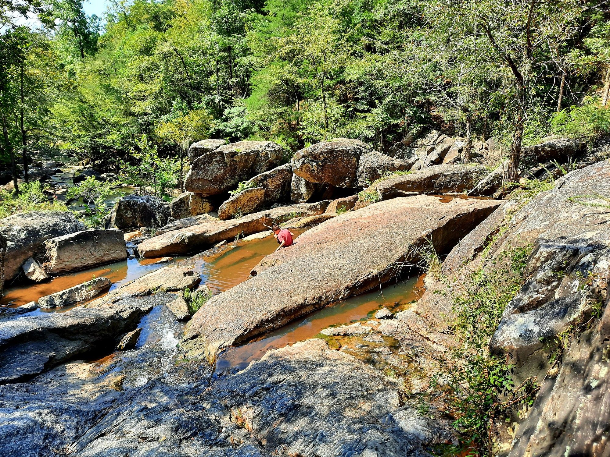 Person in red shirt sitting on large flat rock by a shallow stream. - Chewacla Falls loop from Chewacla State Park