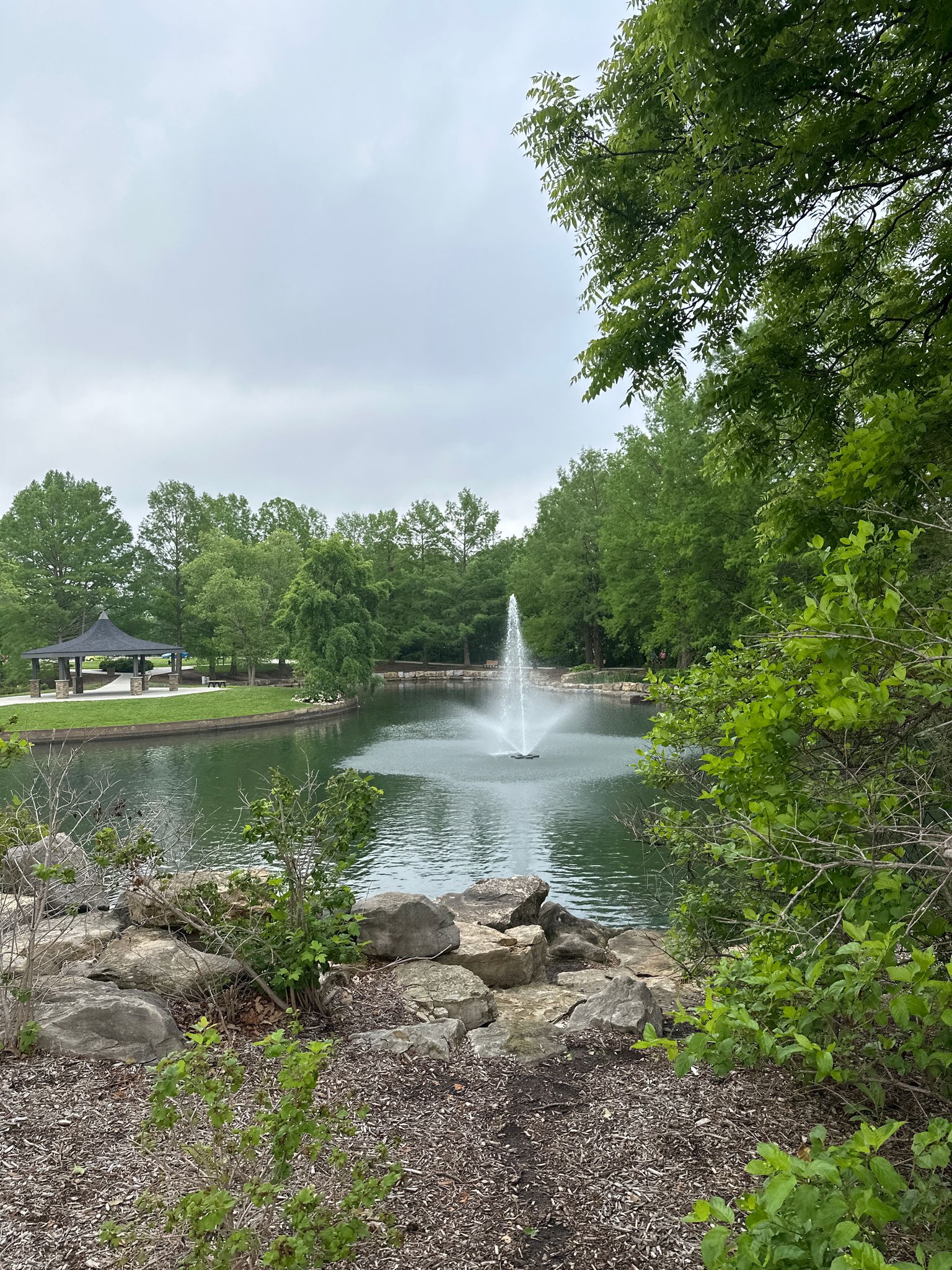 A serene pond with a fountain, lush green trees, and an overcast sky. - City Centre Trail