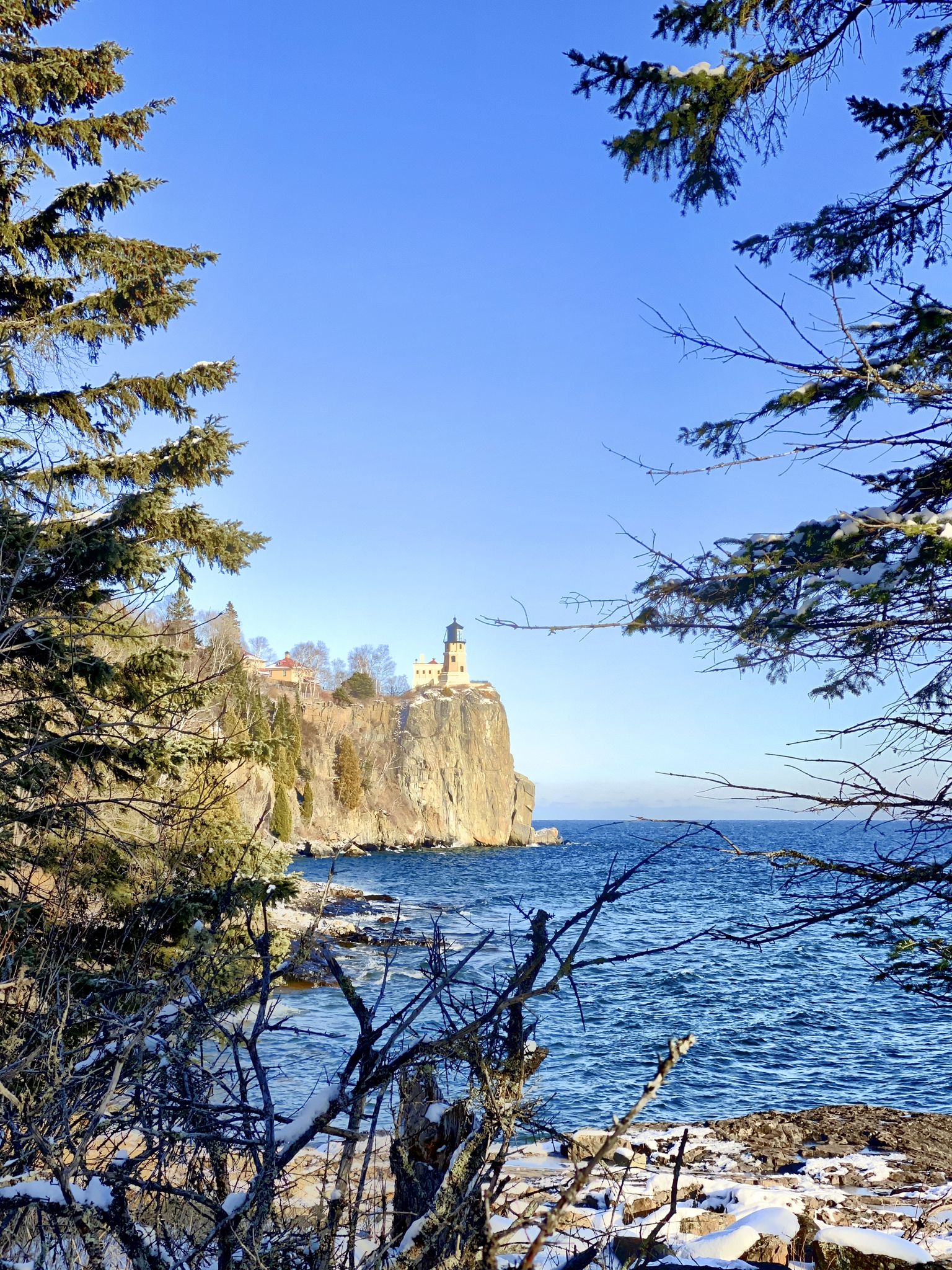 Distant lighthouse on a cliff, framed by snowy evergreen trees and blue water. - Little Two Harbors Trail