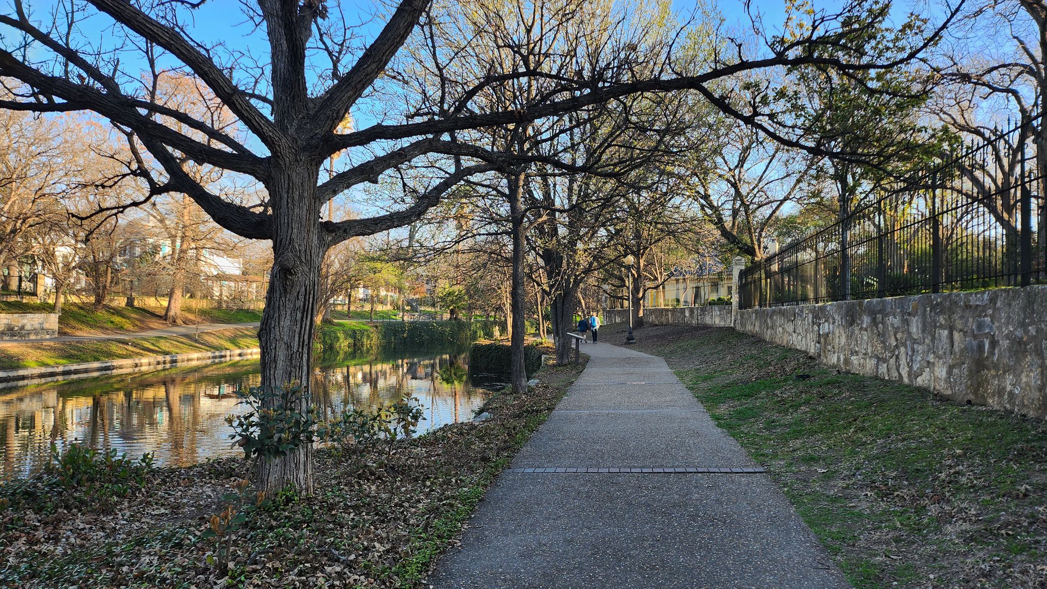 Paved path alongside a calm river with bare trees and distant walkers. - River Walk – San Antonio River Walk loop from Trinity University