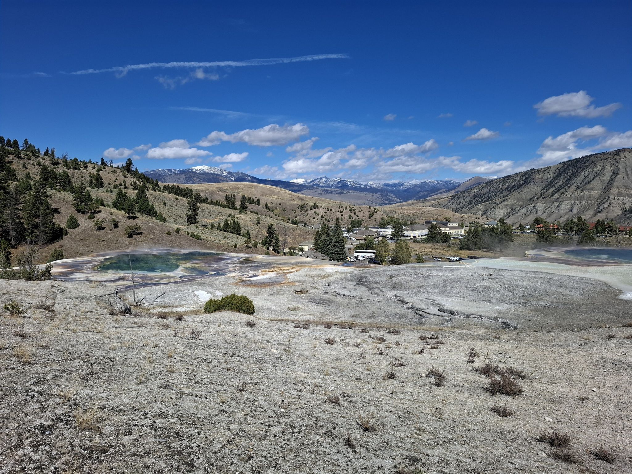 Expansive view of colorful hot springs, hills, and distant mountains under a blue sky. - Mammoth Terraces Trail