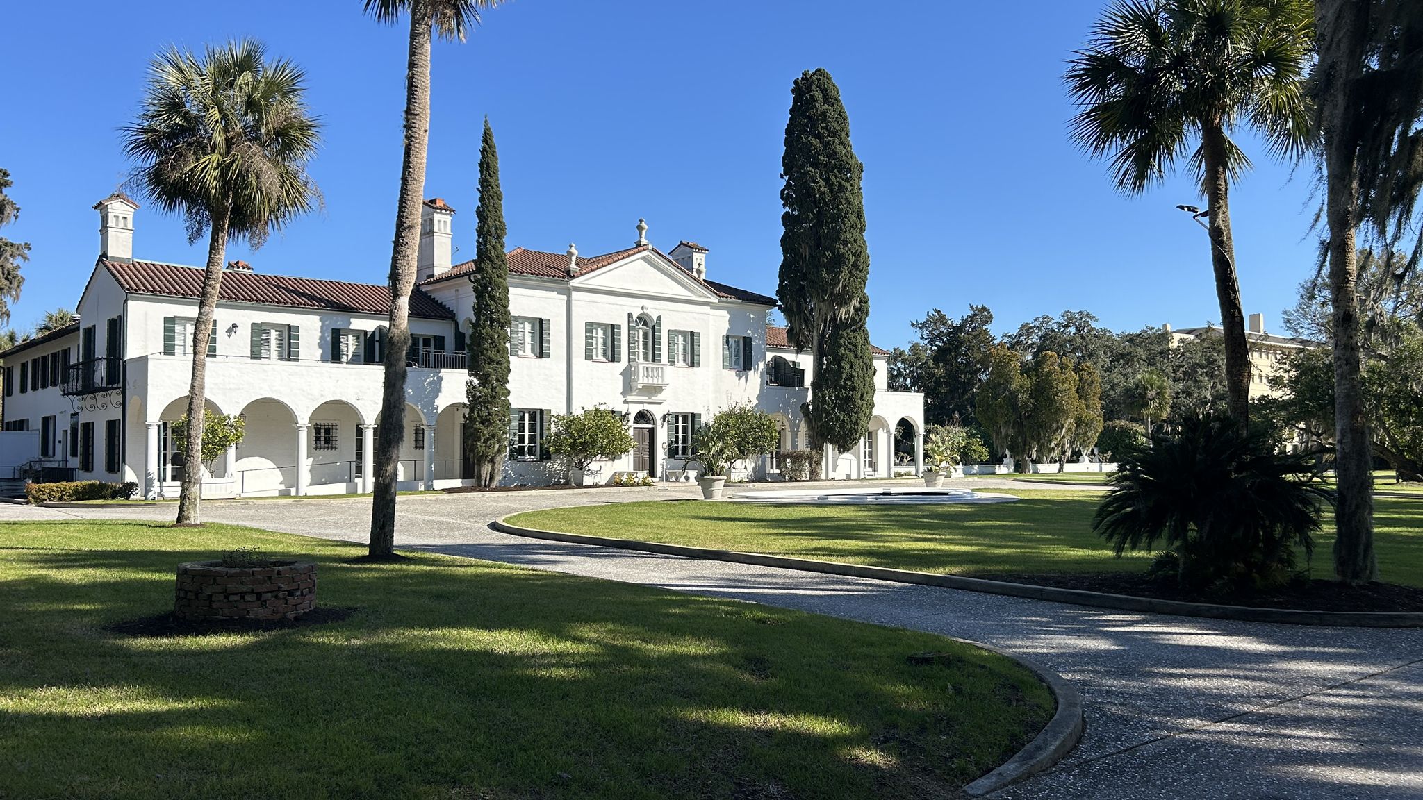 A grand white mansion with a circular driveway, green lawn, and palm trees. - Driftwood Beach – Jekyll Island Clubhouse loop from Jekyll Island
