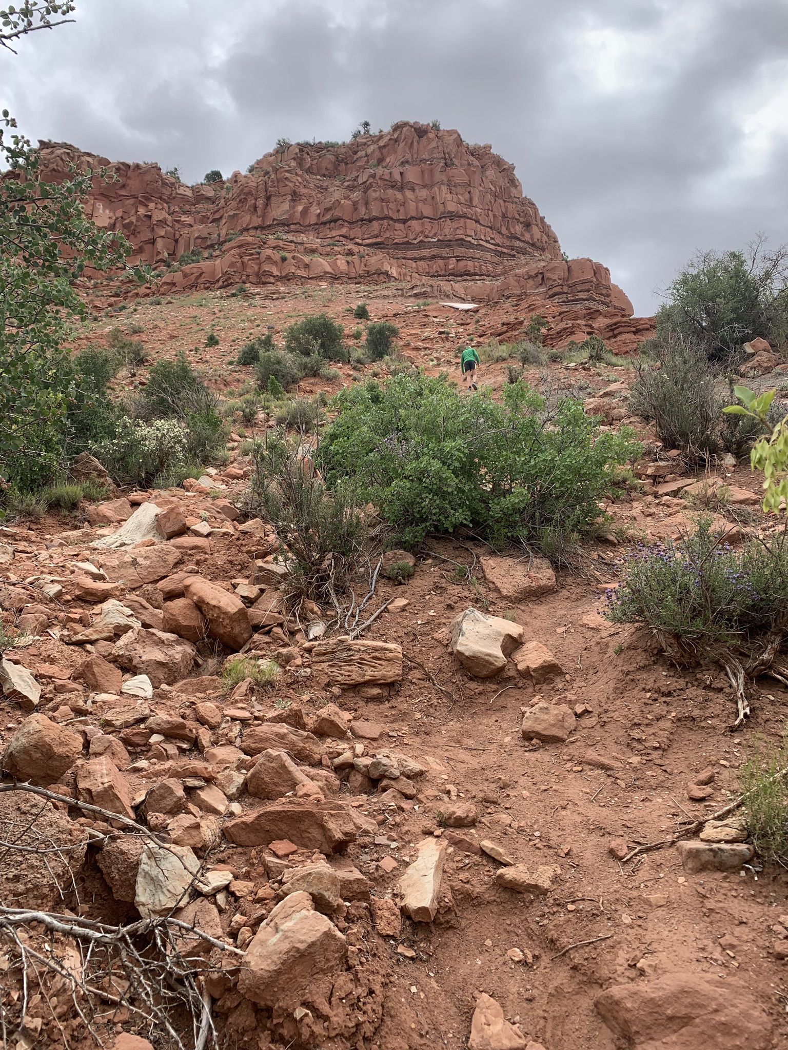 Hiker ascends a rocky red dirt trail towards a large red mountain. - K-Hill – K-Hill Trail loop from Kanab