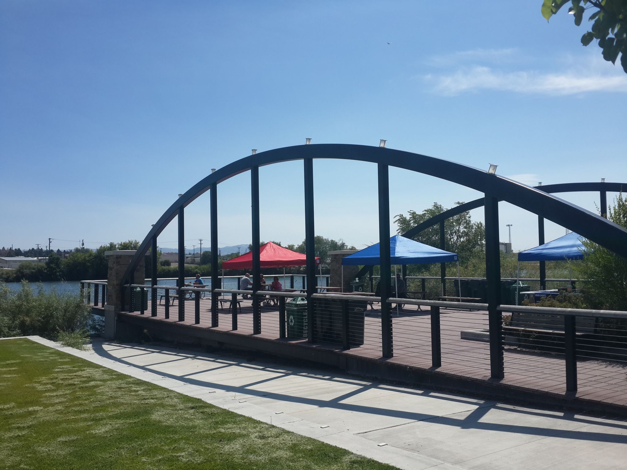 Arched metal bridge and wooden deck with colorful umbrellas on a sunny day. - Idaho Falls Pier loop from Idaho Falls