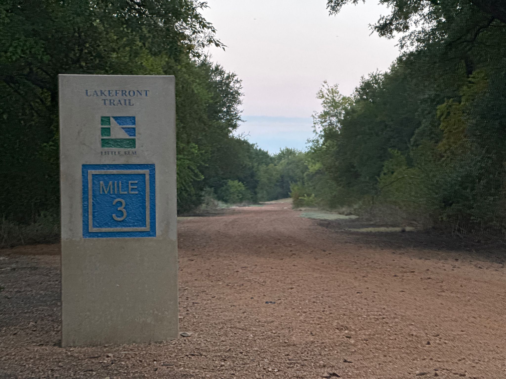 Concrete mile marker 3 beside a dirt trail through dense green trees. - McCord Park — Gravel section – Little Elm Lakefront Trail loop from Bank of America