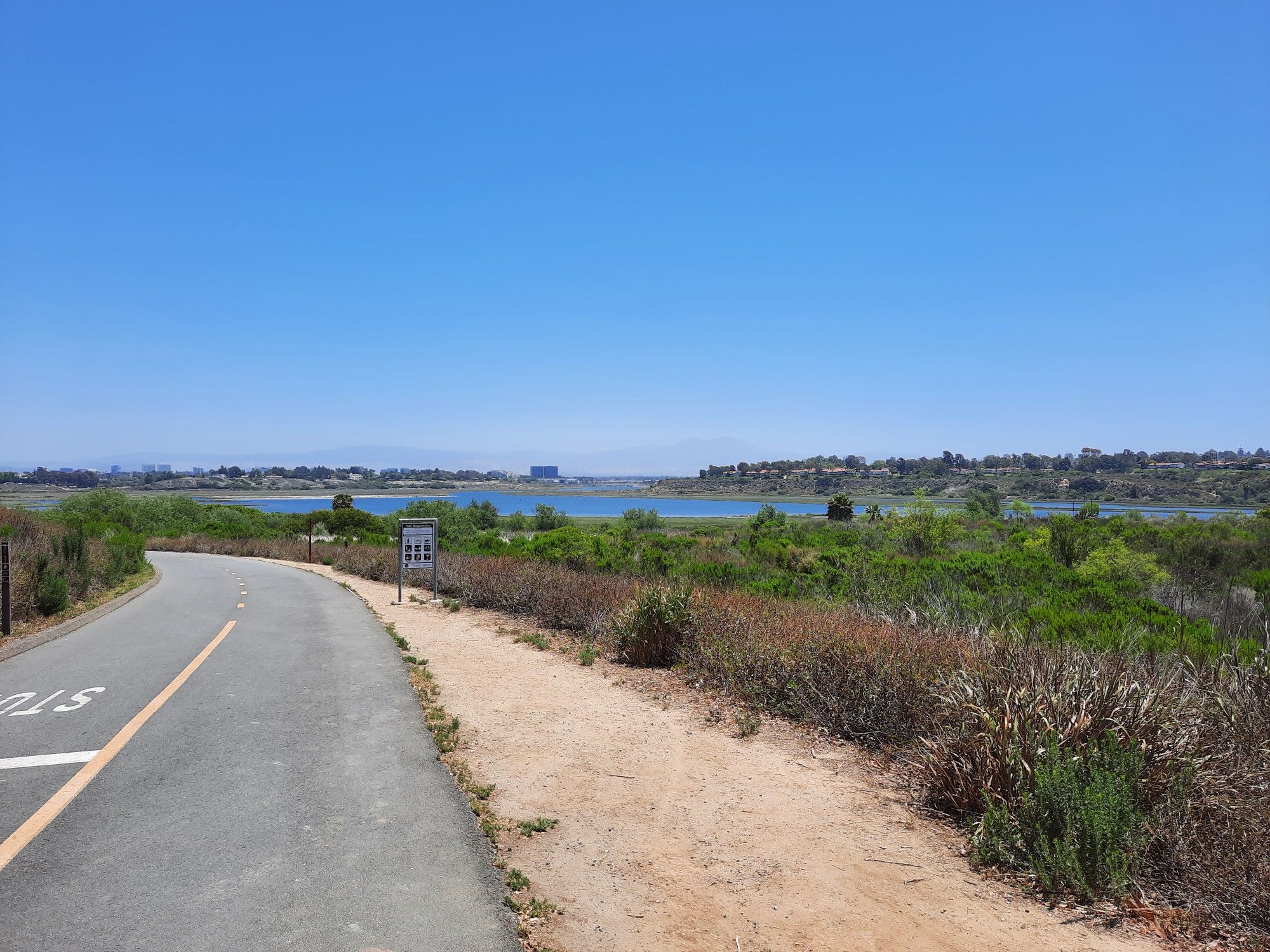 Paved road and dirt path curving alongside a wide wetland under a clear sky. - San Diego Creek views loop from Costa Mesa