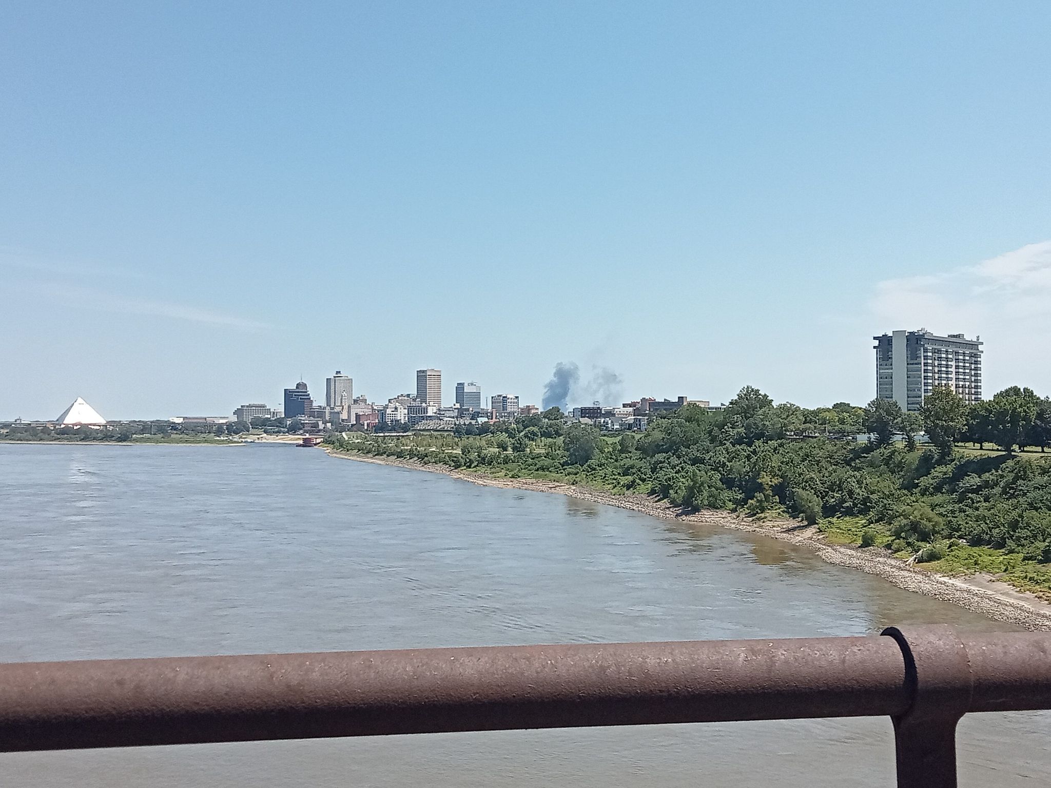 River with a distant city skyline, smoke plume, and a foreground railing. - Big River Crossing loop from FedExForum