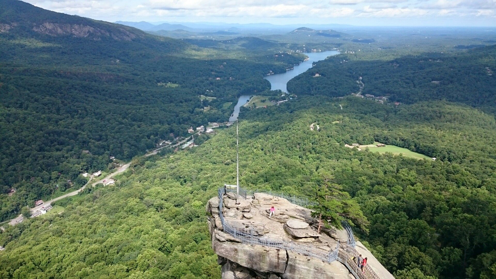 Aerial view of a rocky peak with a flag, overlooking a winding river and forest. Bright daylight, people on peak. - Exclamation Point Trail