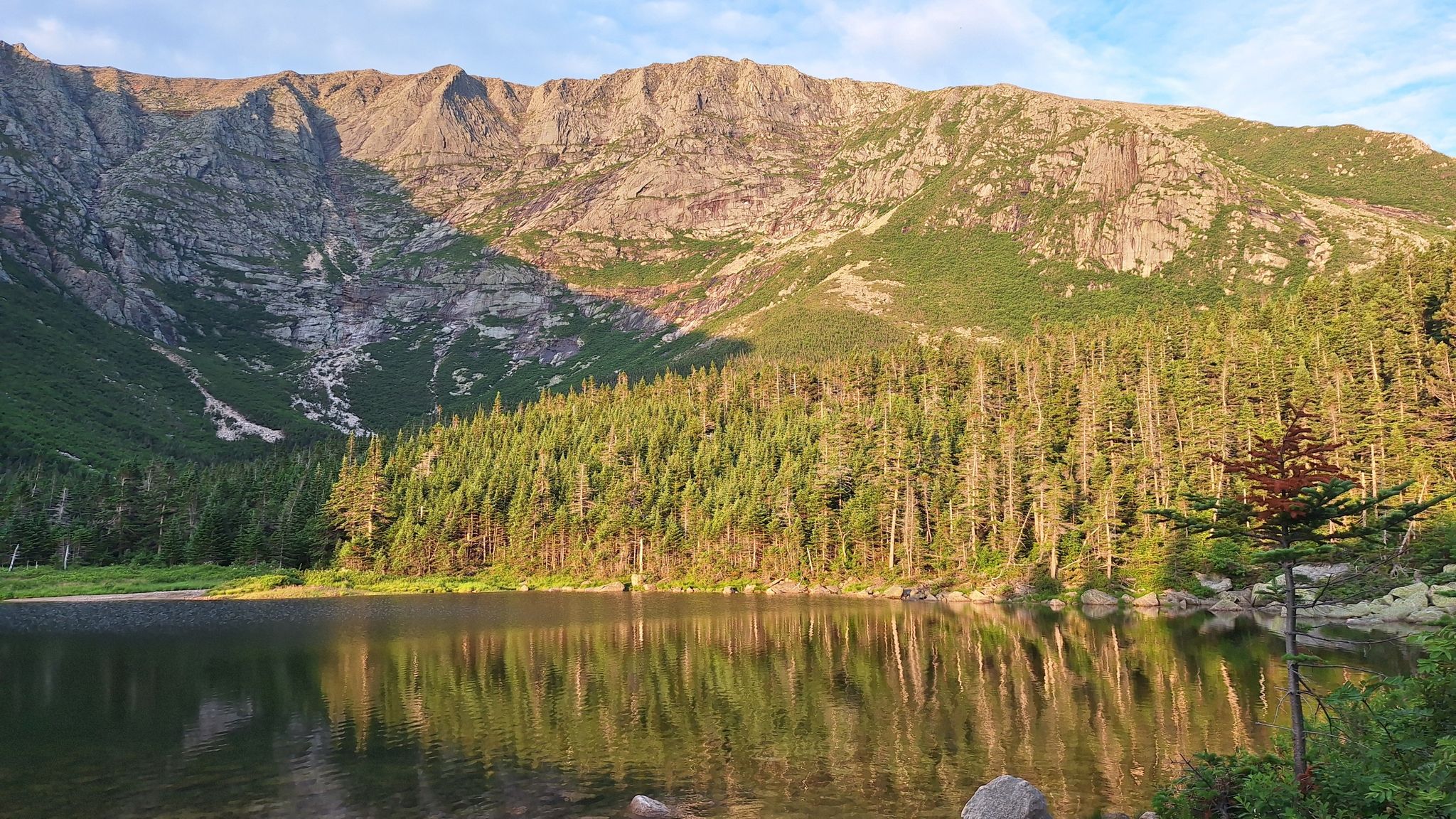 Calm lake reflecting sunlit forest and a towering rocky mountain face. - Chimney Pond Trail