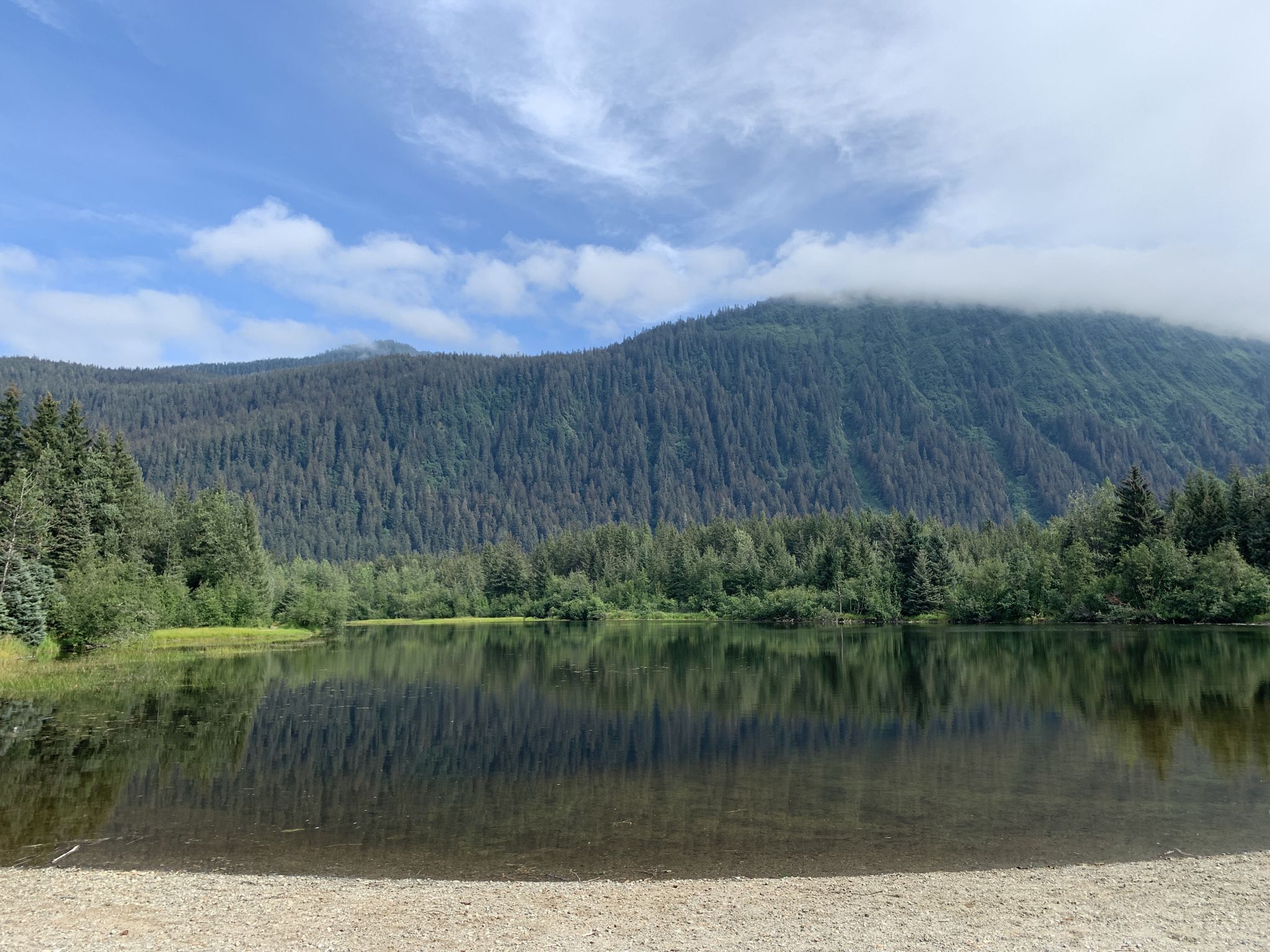 A calm lake reflects forested mountains under a partly cloudy sky. - Dredge, Crystal, Moraine and Moose Lakes Loop