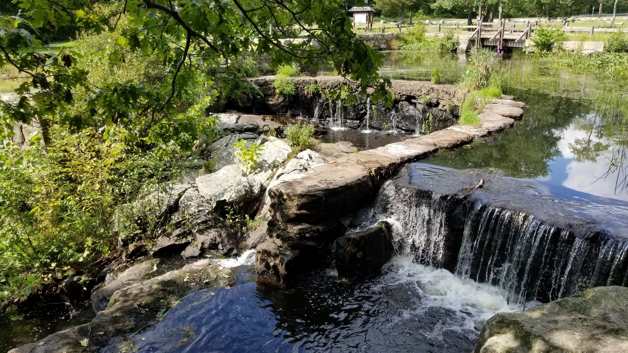 Sunny view of small waterfalls flowing into a pond with a stone path. - Monroe – Southford Falls loop from Old Mine Park