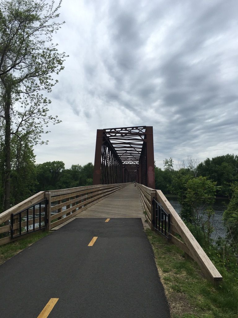 Paved path leading to a large metal truss bridge over a river, cloudy sky. - Norwottuck Rail Trail Bridge – View of the Connecticut River loop from Northampton
