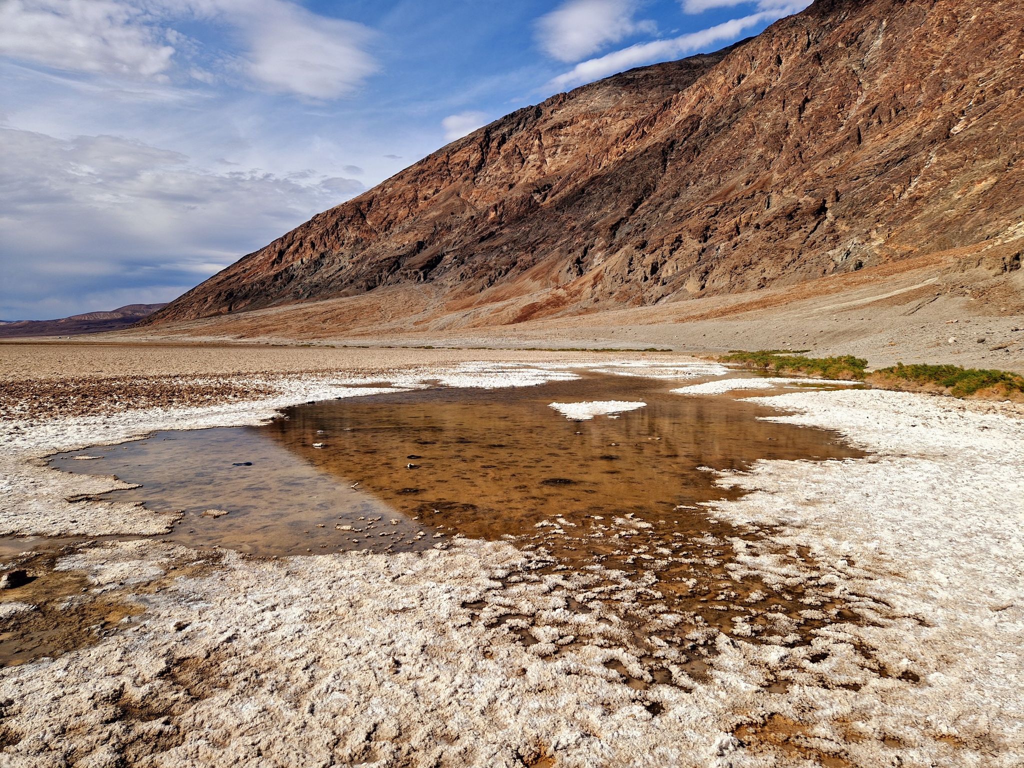 A shallow pool of water on a white salt flat with mountains under cloudy sky. - Badwater Basin Salt Flats Trail