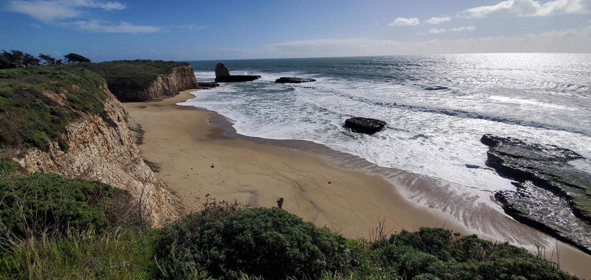 Sunny beach with cliffs, a sea stack, and a person walking by the ocean. - Shark Fin Cove Beach – Natural Bridges State Beach loop from Twin Lakes State Beach