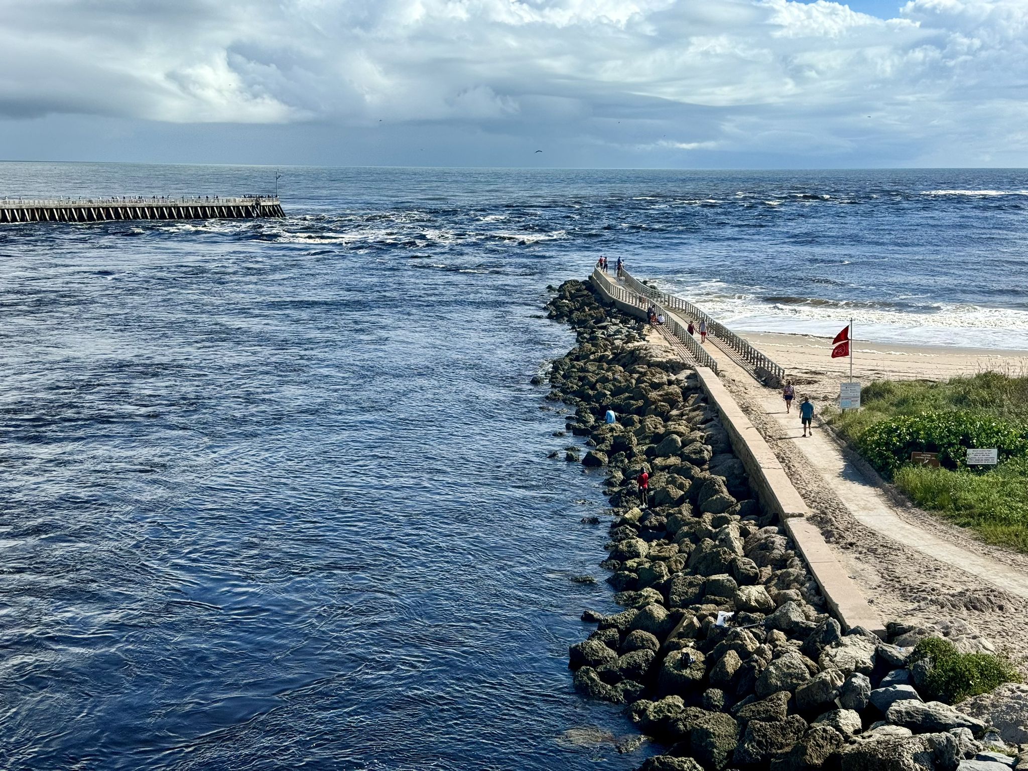 People walk on a concrete jetty separating calm and wavy ocean waters. - Indian River Lagoon Loop via East Coast Greenway