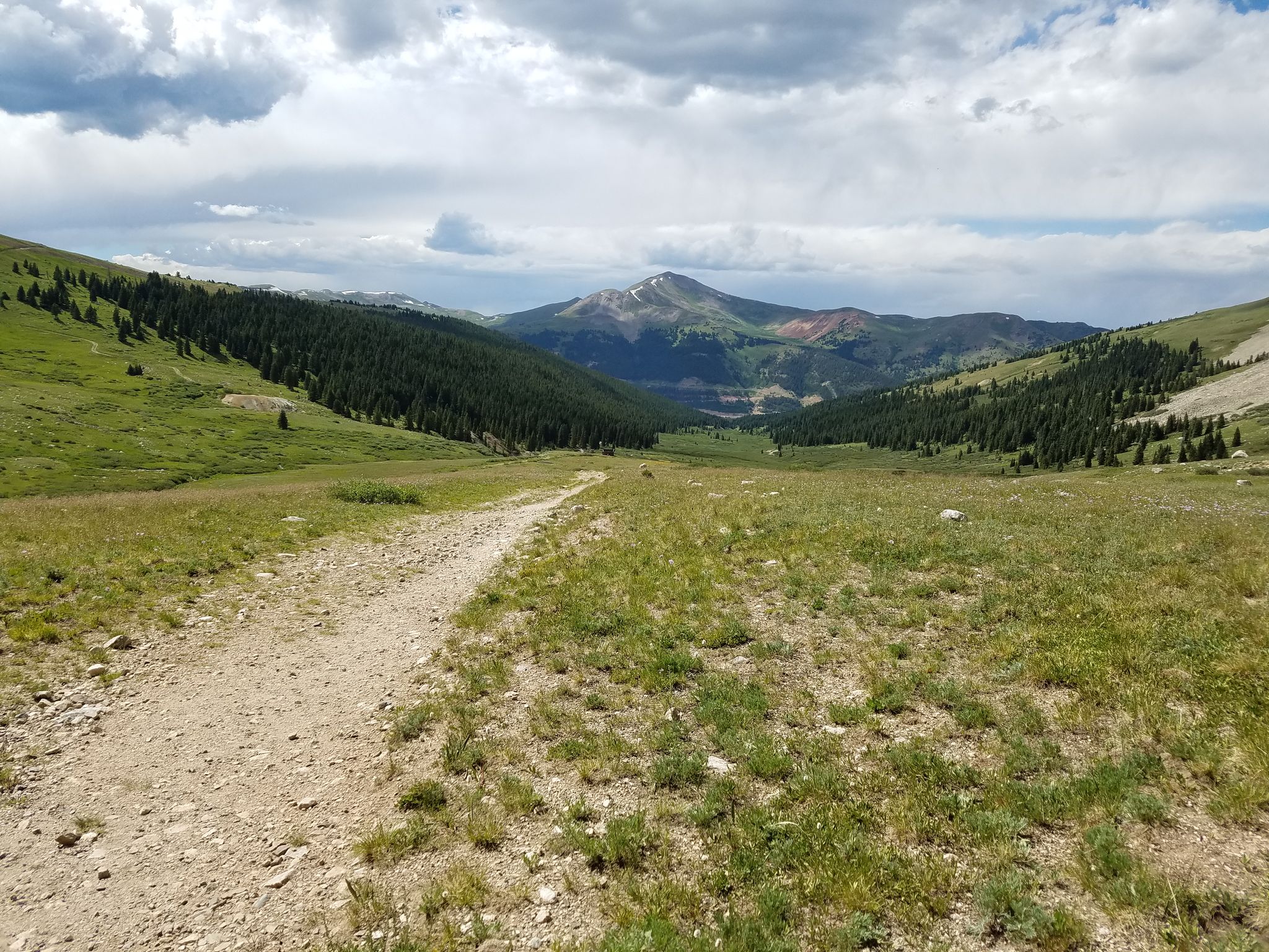 Dirt path winding through a green mountain valley under cloudy skies. - Mayflower Gulch Trail