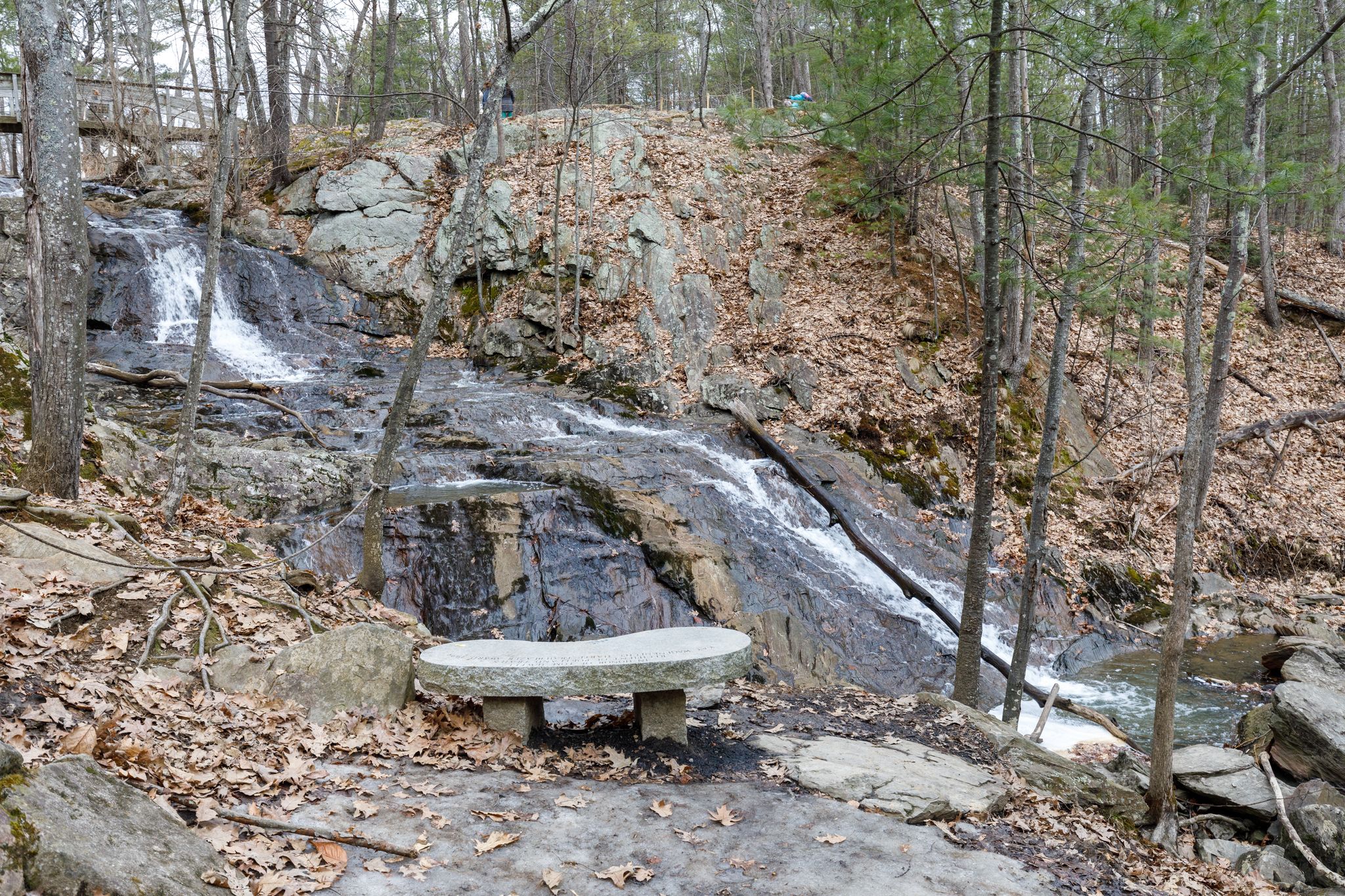 A stone bench overlooks a rocky waterfall in a bare, wooded landscape. - Jewell Falls via Rowe Avenue